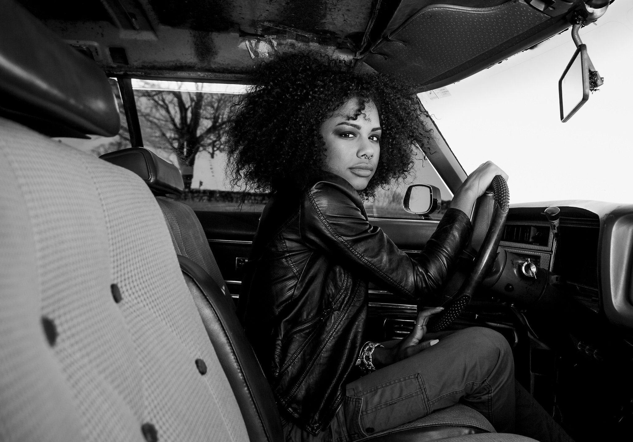 A woman with curly hair and a septum piercing sits in the driver's seat of a car, looking at the camera. The photo is in black and white, and she is wearing a leather jacket.