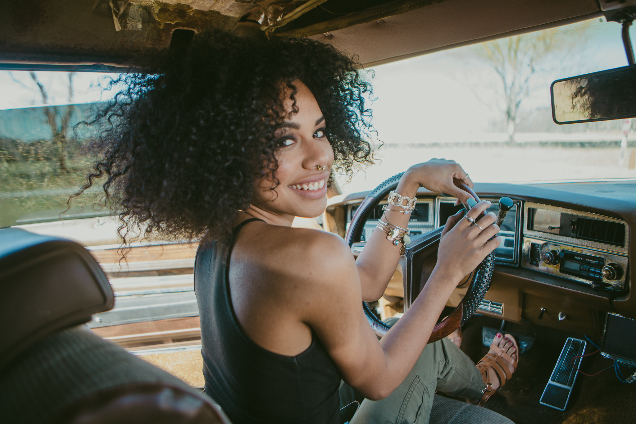 A woman with curly black hair smiling while sitting in the driver's seat of a vintage vehicle, with her left hand on the steering wheel. She is wearing multiple rings and bracelets, and beige sandals. The background shows trees and a clear sky throug