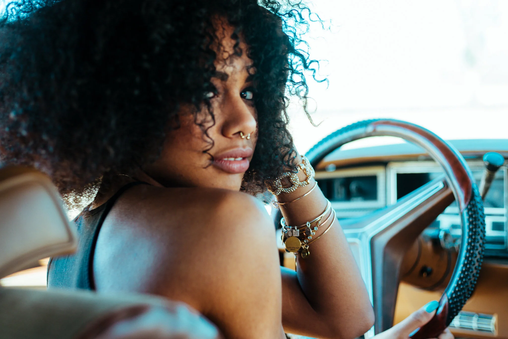 Close-up of a young woman with curly hair and multiple nose piercings, sitting in a vintage car, wearing jewelry, and looking over her shoulder.