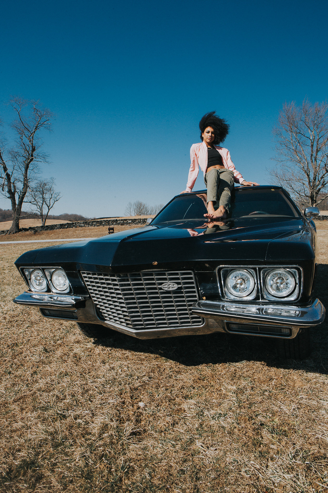 A woman with curly black hair sitting on the hood of a vintage black car in an open field with leafless trees, under a clear blue sky.