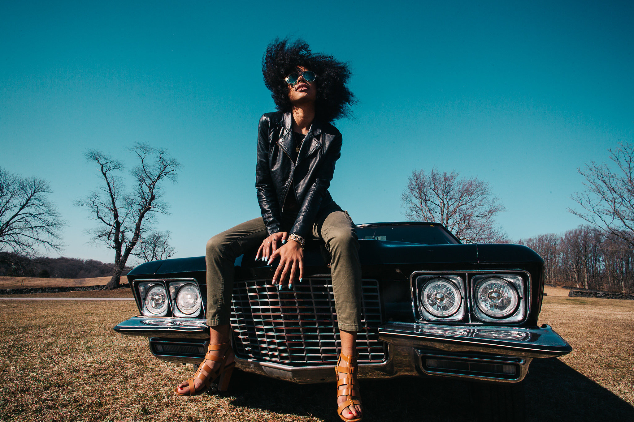 A woman with curly hair wearing sunglasses, leather jacket, and tan sandals sitting on the hood of a classic black Cadillac car in an open field with bare trees and clear blue sky in the background.
