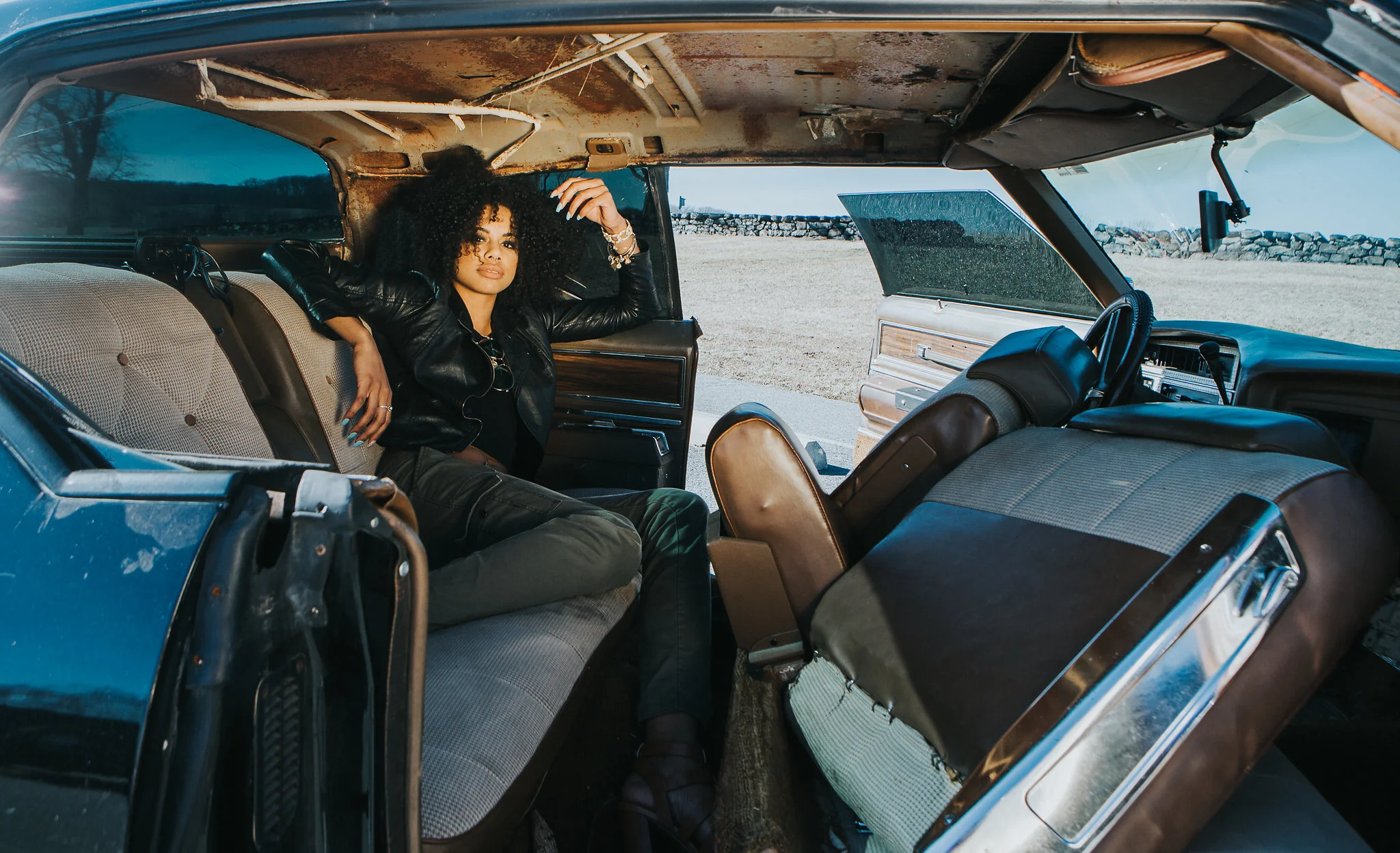 A woman with curly black hair sitting inside an old, partially disassembled car, outdoors near a stone wall and grassy field, wearing a black leather jacket and jewelry.