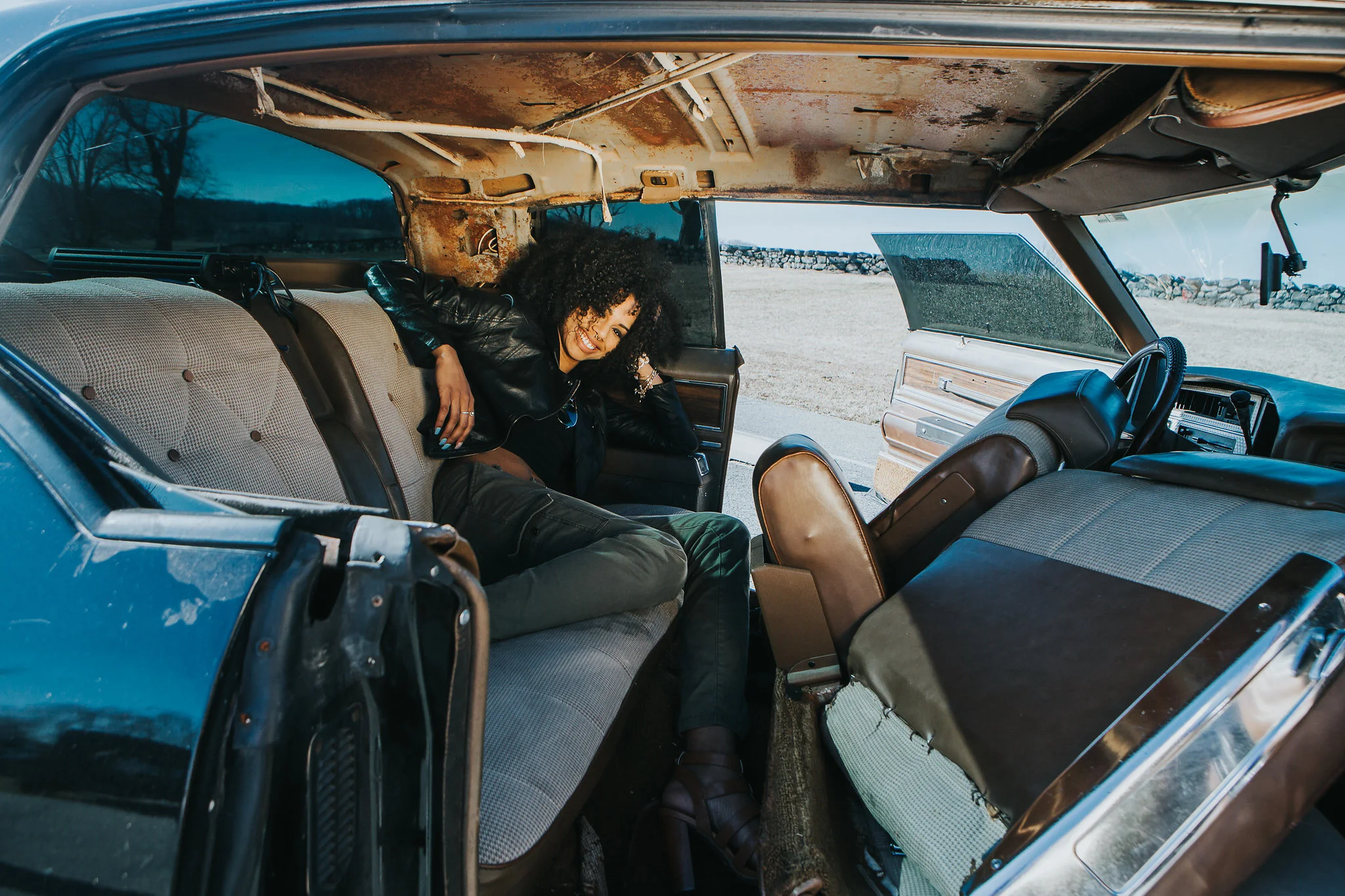 A woman with curly hair and a black leather jacket smiling inside an old car with a rusty interior, parked outdoors near a rural landscape.