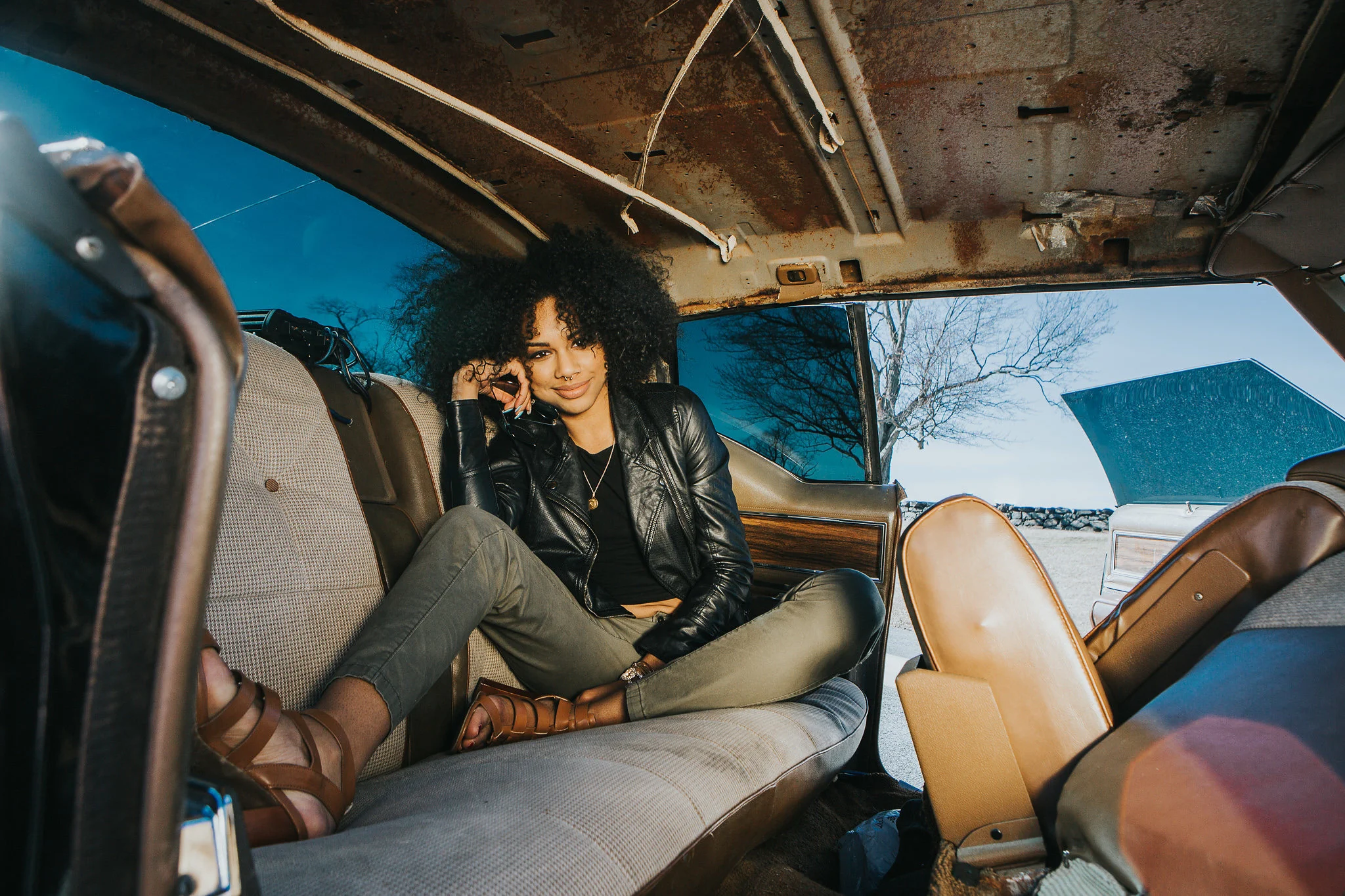 A woman with curly hair and a leather jacket sitting inside an old, rusty vehicle, looking at the camera with a smile, and holding a phone while wearing sandals and casual pants.
