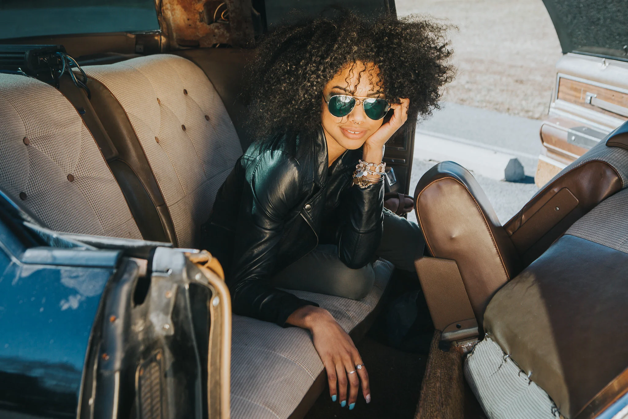 A woman with curly hair wearing sunglasses, a black leather jacket, and multiple bracelets, sitting inside a vintage vehicle, possibly an RV or bus, with a scenic outdoor background.