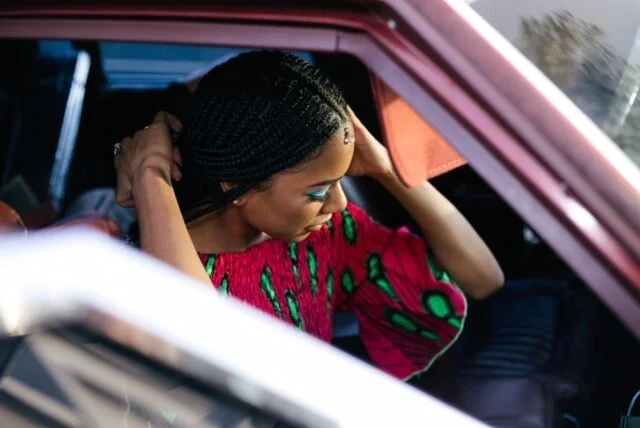A woman with braided hair and bright makeup adjusting her hair in a pink vehicle, wearing a colorful patterned top.