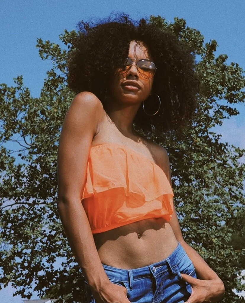 A woman with curly hair wearing sunglasses, hoop earrings, an orange strapless top, and blue jeans standing outdoors under a tree with a blue sky background.