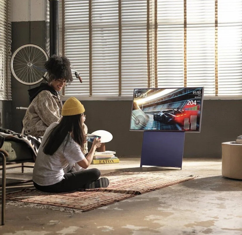 A woman and a girl sitting on a rug in a room, watching a racing video game on a TV. The girl is holding a white game controller and wearing a yellow beanie, while the woman has curly hair and is smiling. The room features blinds covering large windo