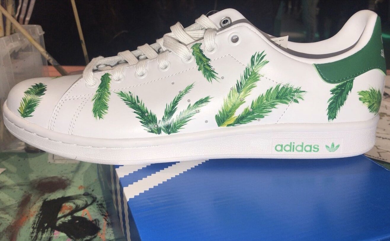 A white Adidas sneaker decorated with green leaf patterns printed on the sides and toe area, resting on a blue shoebox.