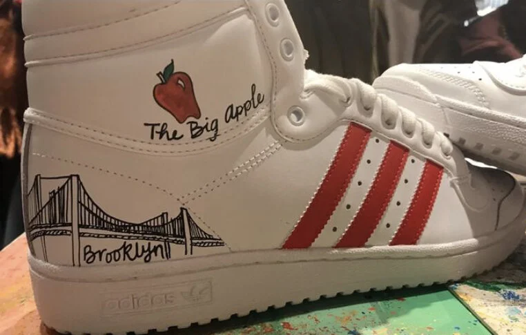 Close-up of a white Adidas high-top sneaker with custom drawings, including a red apple labeled 'The Big Apple,' the Brooklyn Bridge, and red and white stripes.
