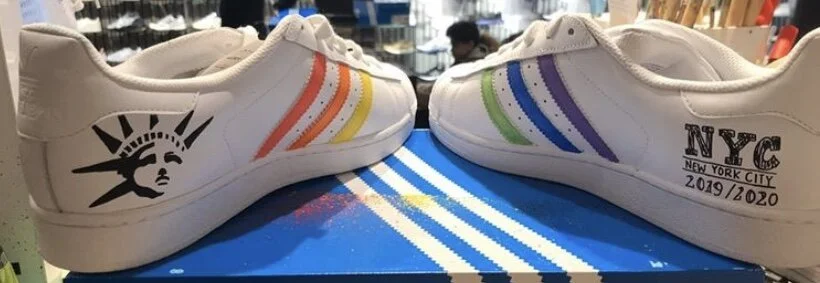 Pair of white sneakers with rainbow stripes, one with an American flag and Statue of Liberty graphic, the other with NYC NYC New York City text and dates 2019/2020, placed on a colorful striped mat.