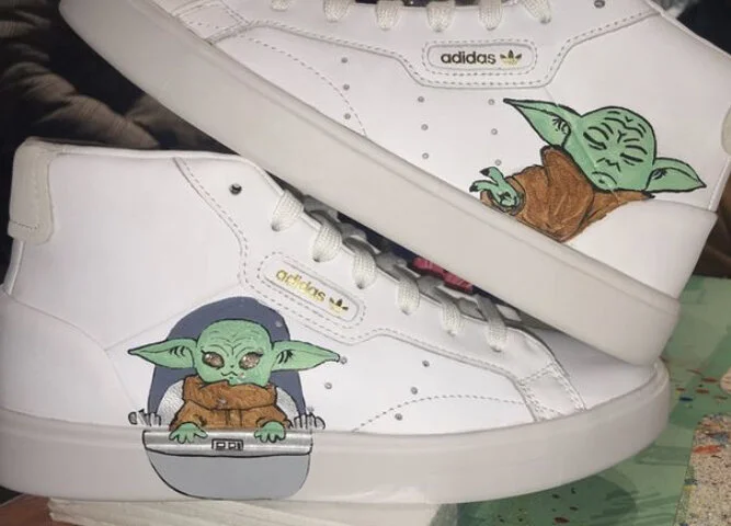 Pair of white sneakers with Baby Yoda (Grogu) cartoon illustrations on the sides; one shoe has Baby Yoda with headphones and a laptop, the other has Baby Yoda in a brown robe with a green head and ears.