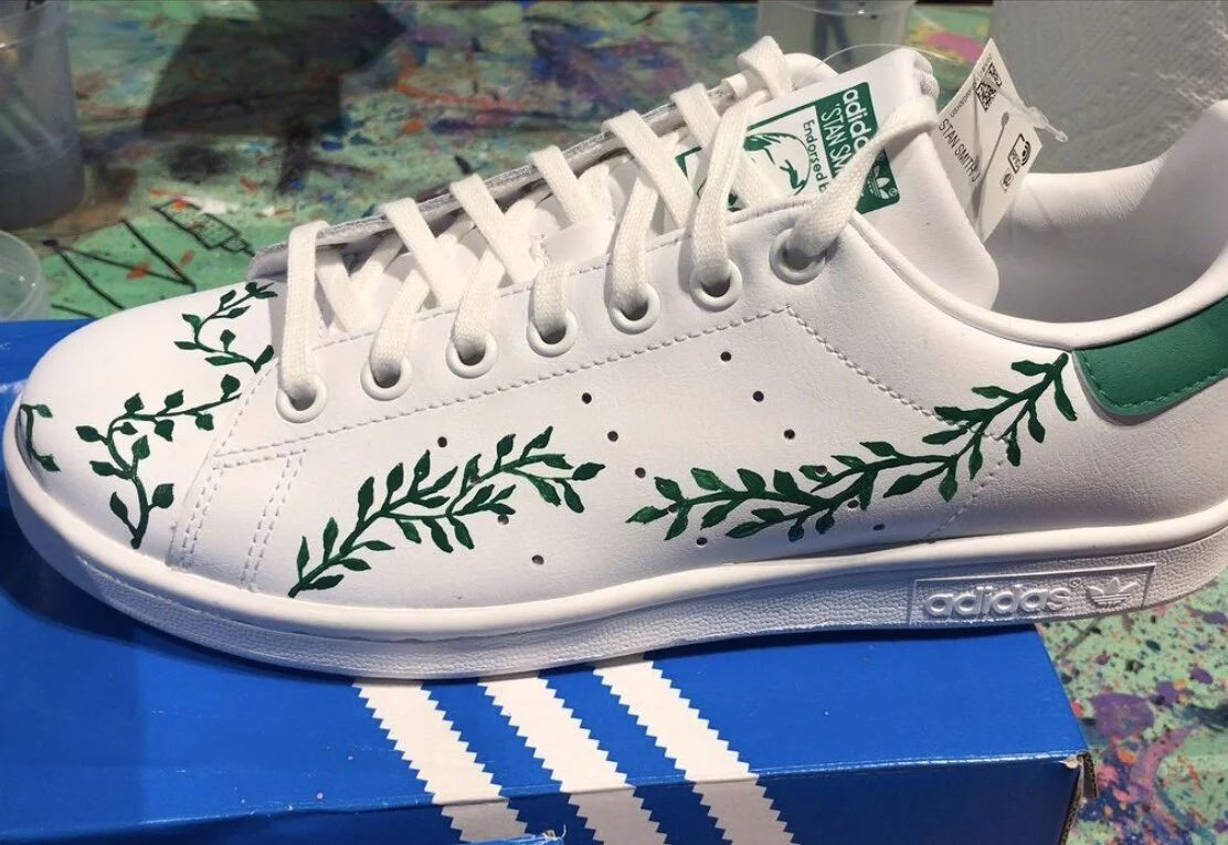 White Adidas Stan Smith sneakers with green leafy vine designs, placed on a blue Adidas shoebox.