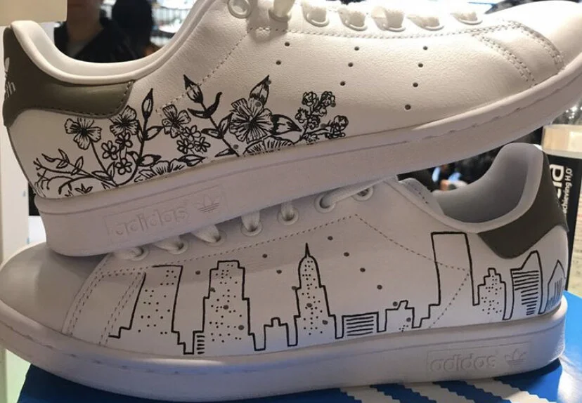 Two white Adidas sneakers with black line drawings of New York City skyline and floral patterns
