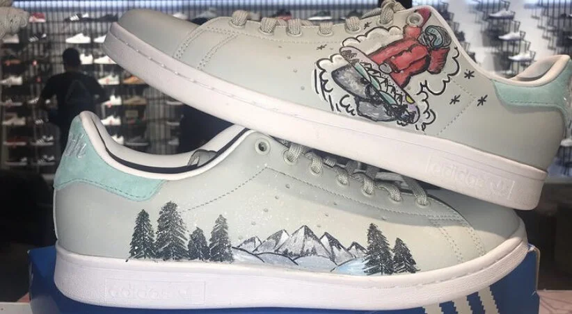 Pair of white Adidas sneakers with hand-drawn mountain landscape and trees on the side, and animated art of a red jacket and skateboard on the top shoe.