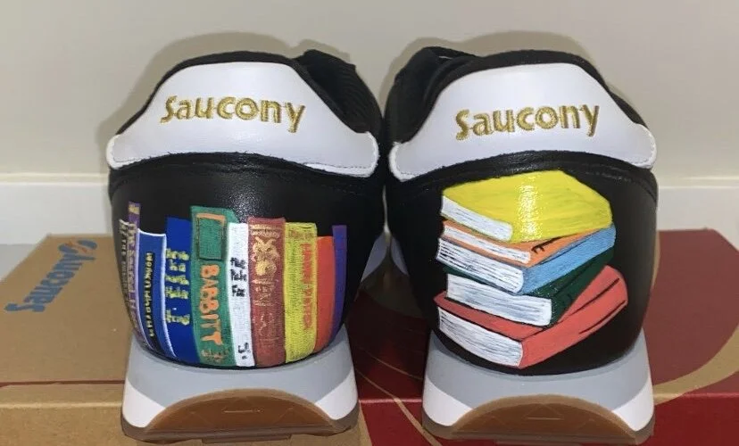 Custom painted Saucony sneakers featuring books on the back, one side with a row of colorful books and the other with a stack of books in various colors.