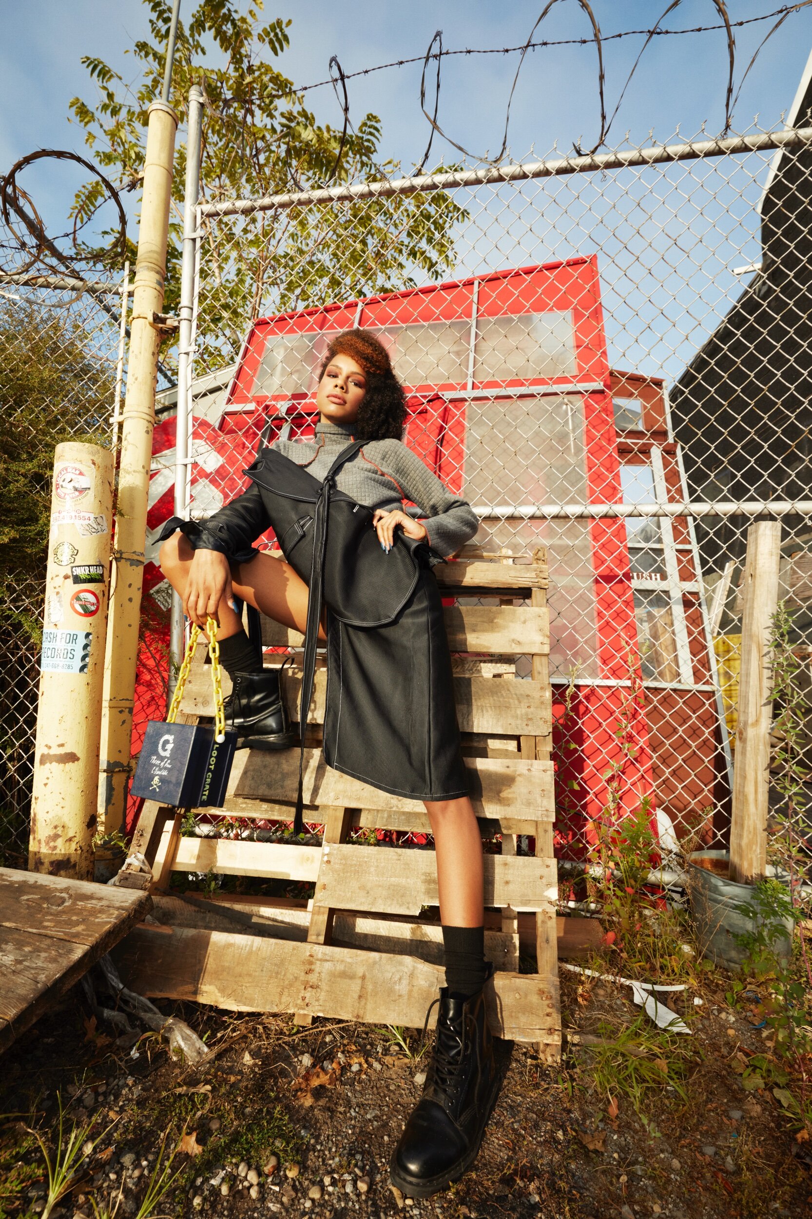 A young woman with curly hair and a serious expression stands confidently in front of a chain-link fence, with a red structure behind her, holding a yellow chain, dressed in layered clothing and black combat boots.