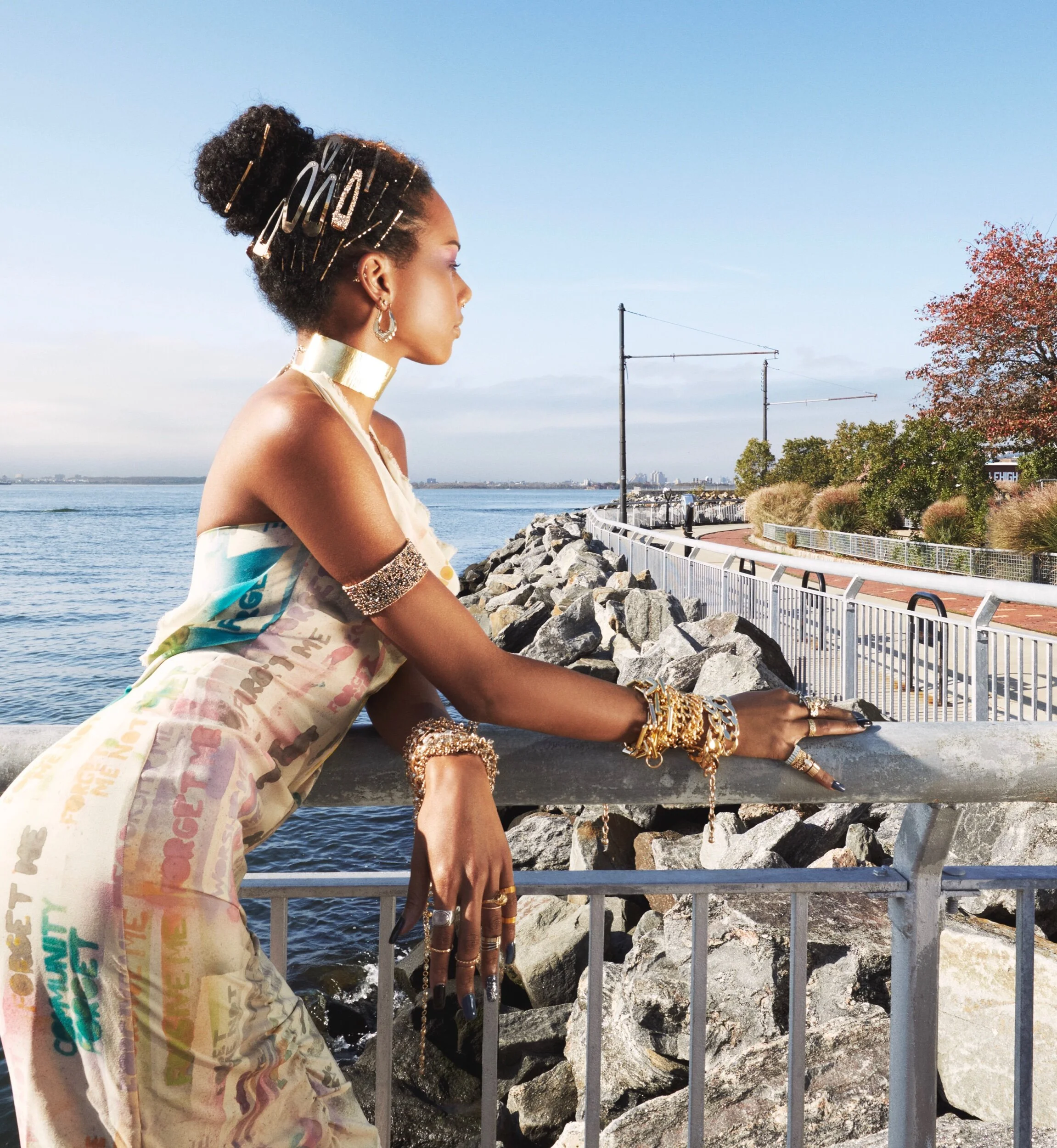 Side profile of a woman with dark hair styled in a large bun, wearing gold accessories and a colorful, patterned off-shoulder top, leaning on a gray stone barrier by a body of water with trees and a clear sky in the background.
