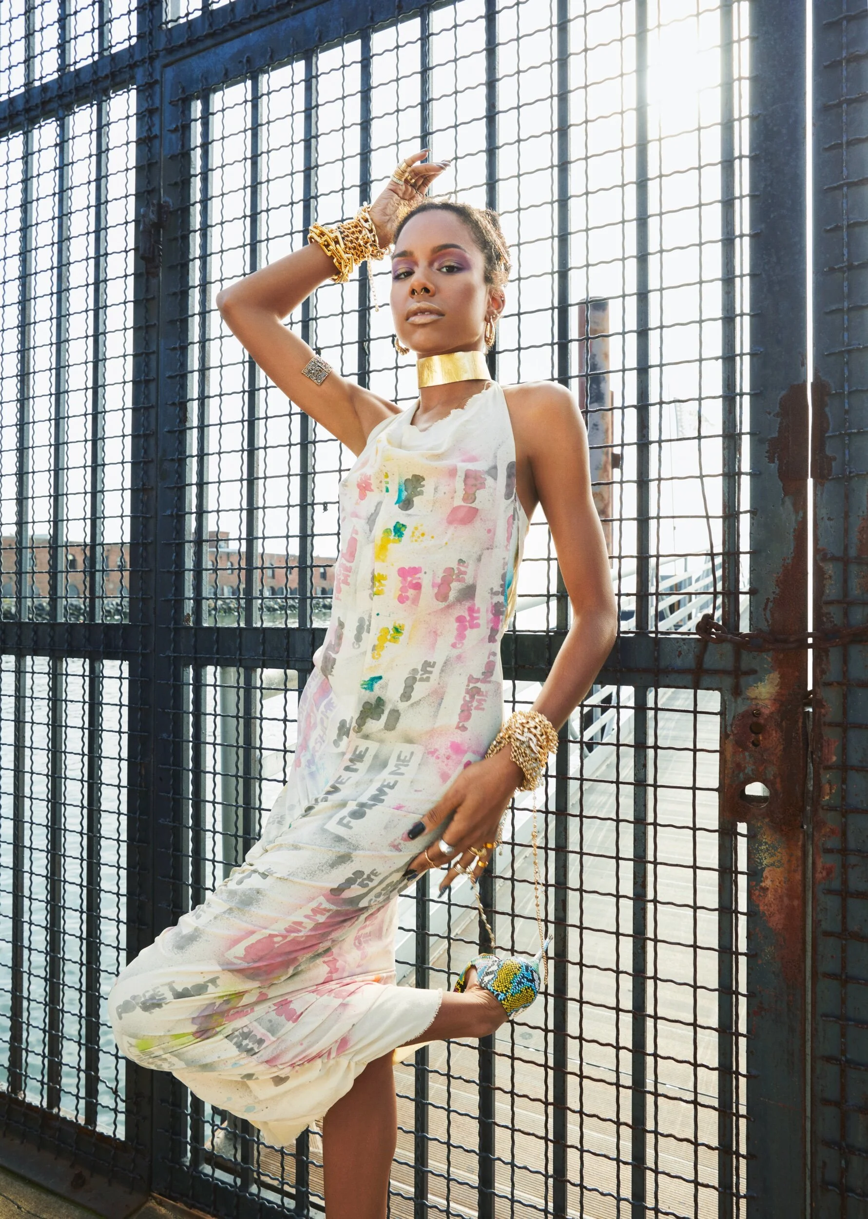 A woman with jewelry and colorful makeup leaning against a metal fence on a city rooftop with a cityscape and sunset in the background.