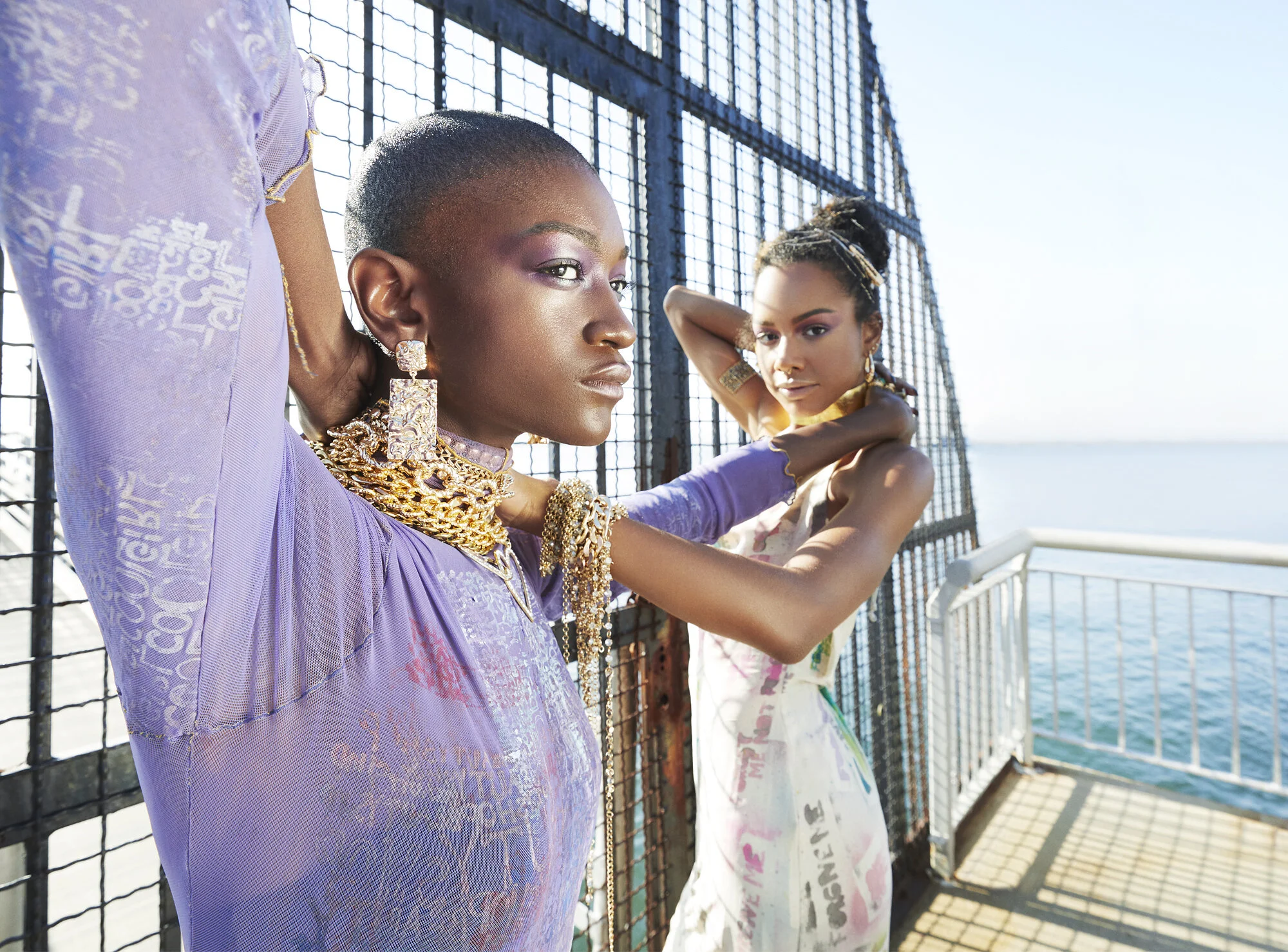 Two women standing strong by a metal fence overlooking the water, wearing colorful, stylish clothing and jewelry, with their arms raised and relaxed poses.