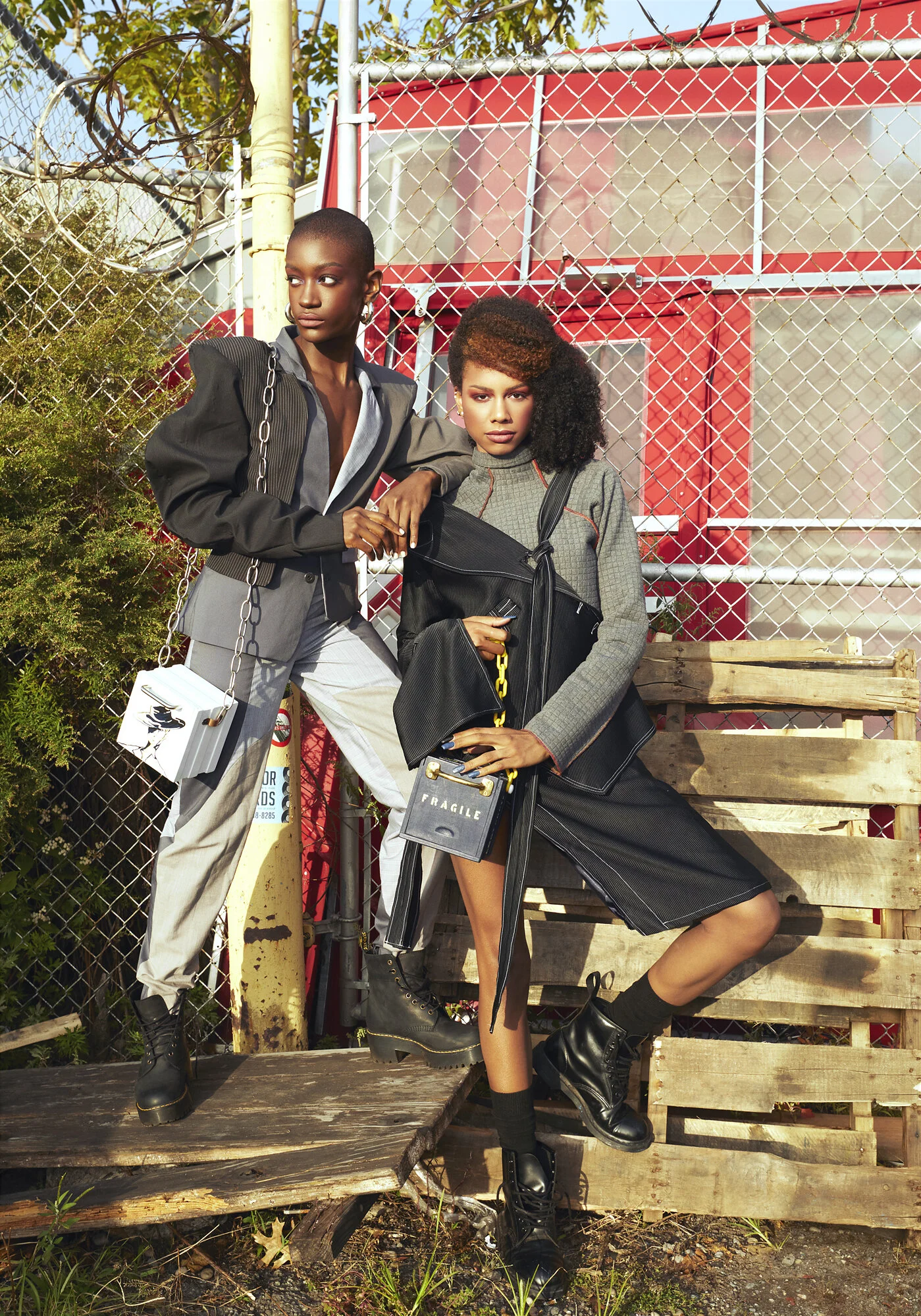 Two women posing fashionably in an outdoor urban setting with a chain-link fence, ladder, and red building in the background. One woman with short hair and hoop earrings leans against the fence, while the other with curly hair and bold makeup stands 