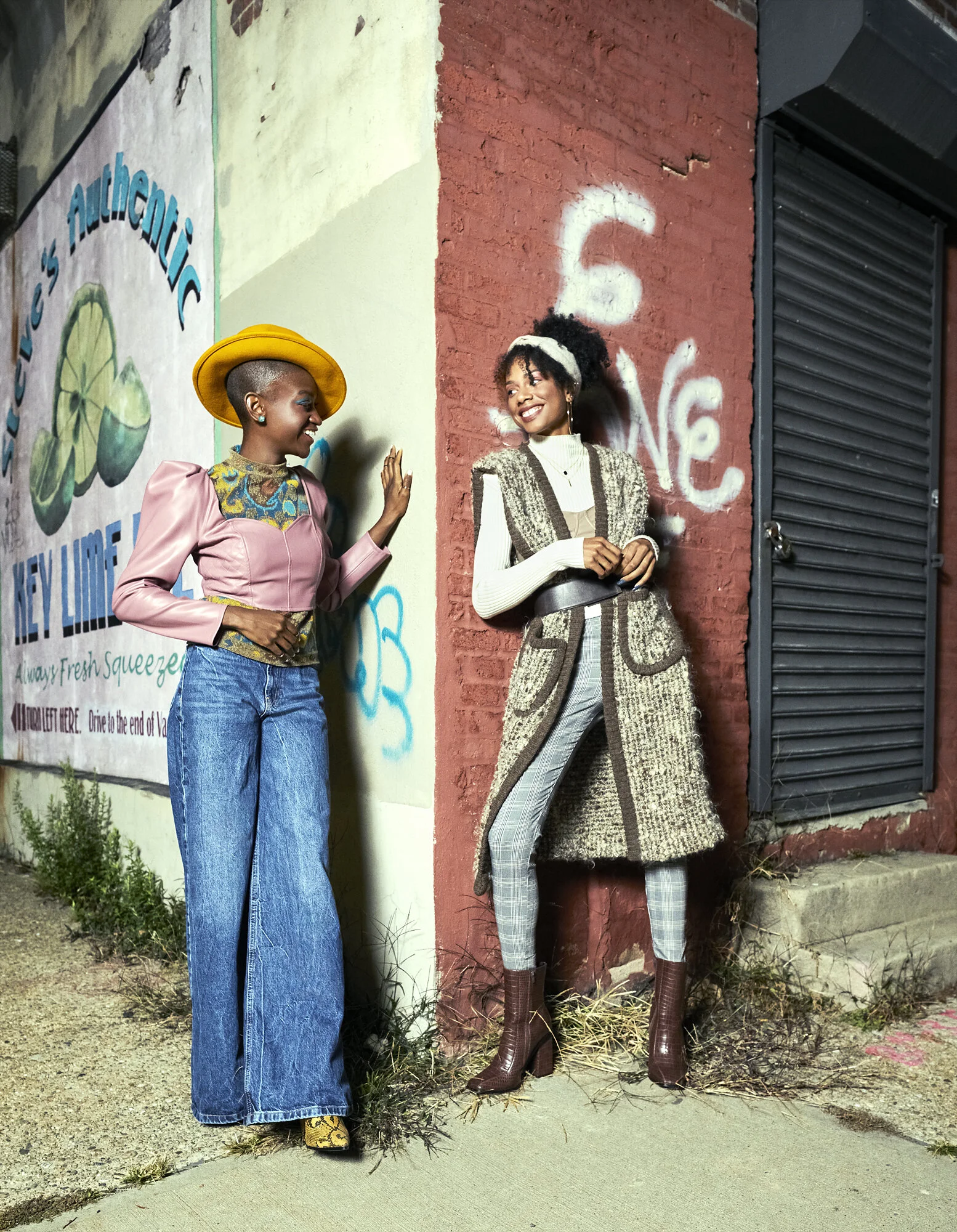 Two women standing on sidewalk near brick and painted walls, smiling and looking at each other. One woman wears a yellow hat, pink jacket, high-waisted blue jeans, and snakeskin boots. The other woman wears a headband, patterned coat, plaid pants, an