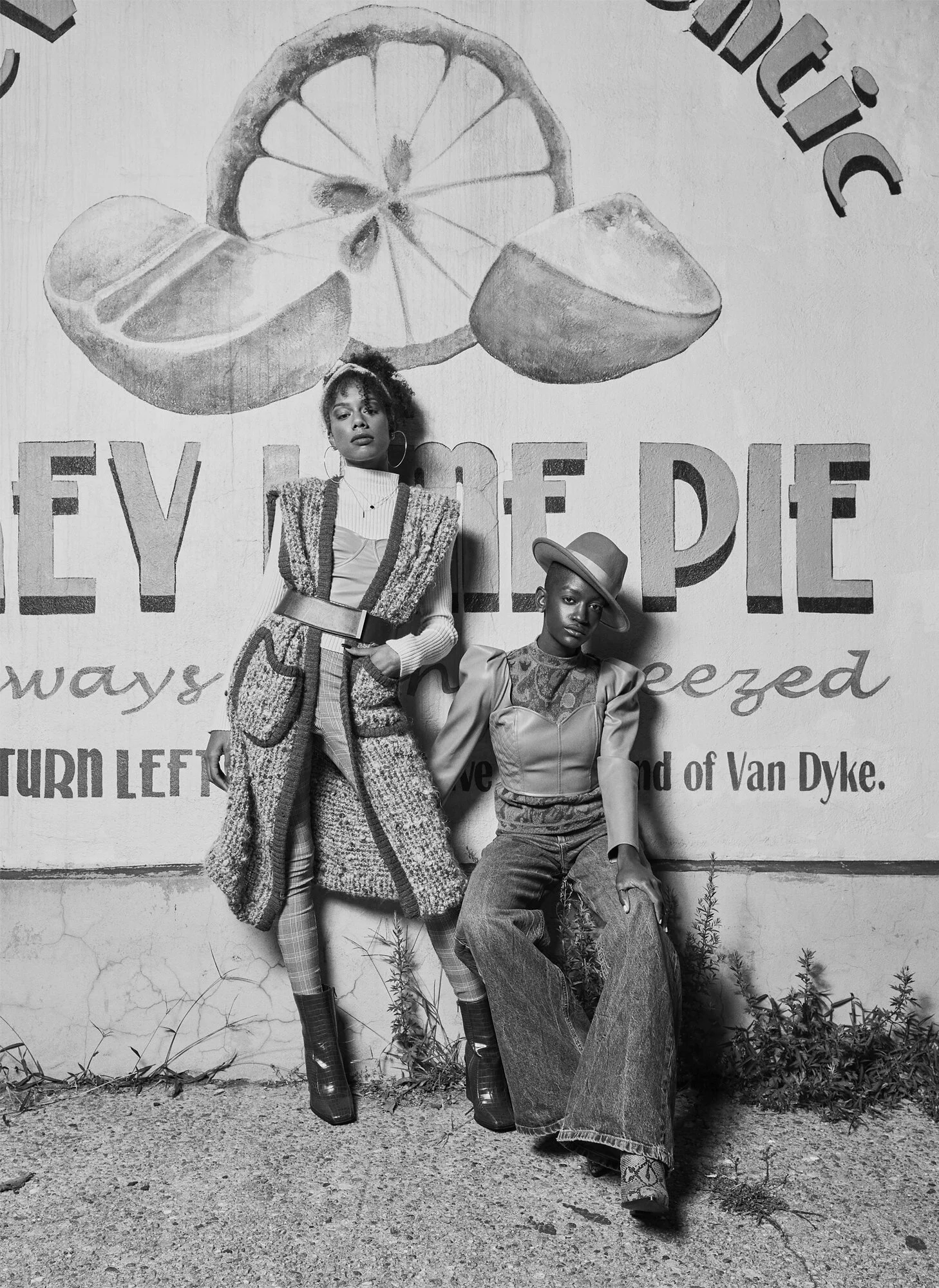Two women with short hair and unique outfits in front of a wall with citrus fruit illustrations and partial text that says "LIFE OF."