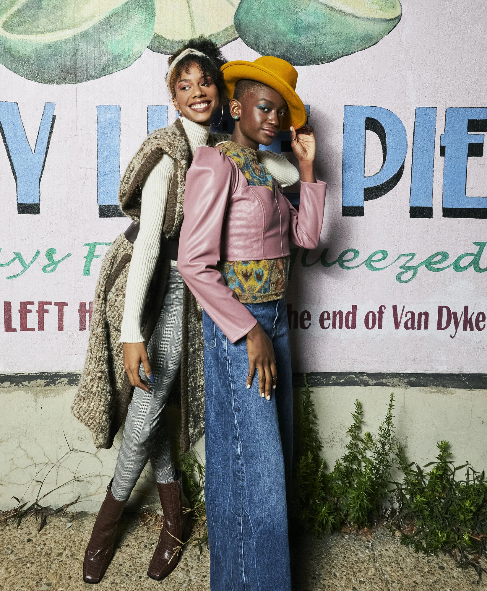Two women standing in front of a painted wall with large green leaves and blue letters. One woman is smiling, wearing plaid pants, a cream turtleneck, a fuzzy vest, and tall brown boots. The other woman, with a serious expression, is wearing a yellow