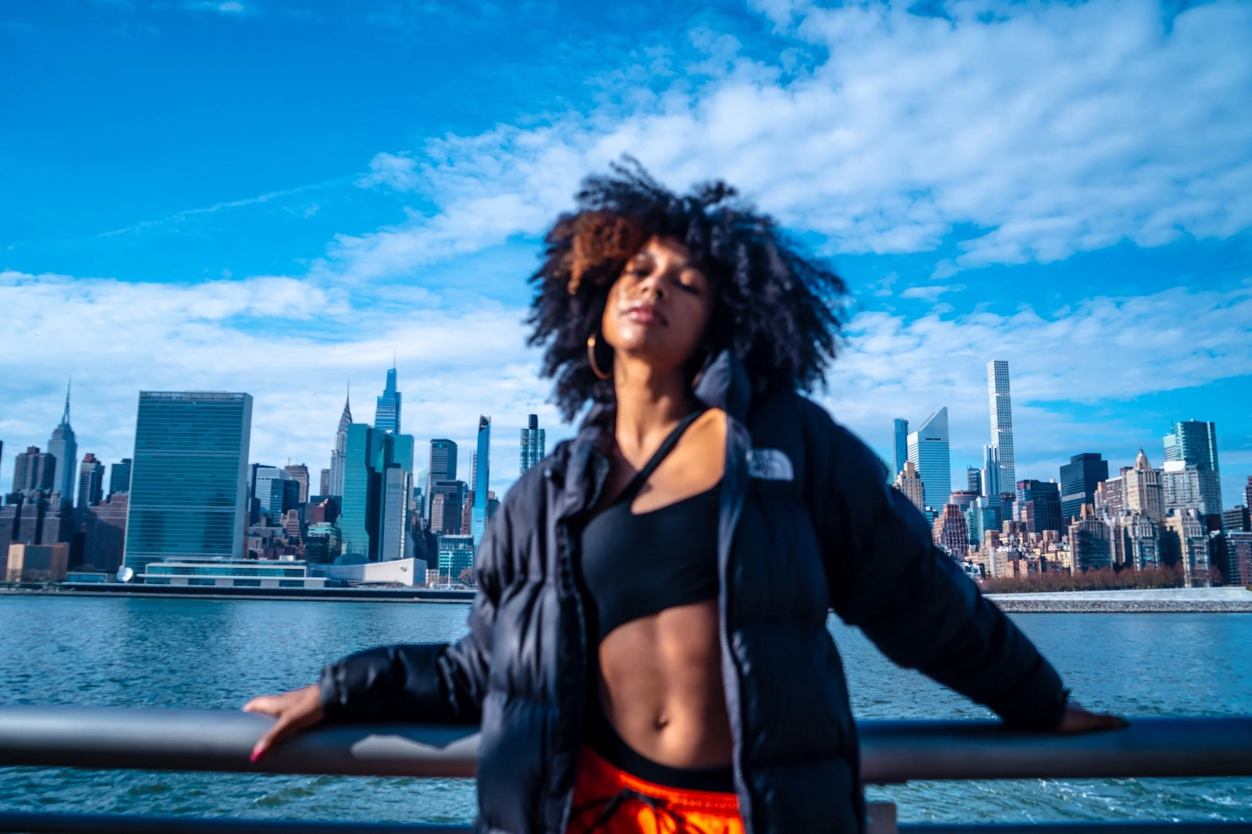 A woman with curly hair standing on a boat with New York City skyline in the background on a clear day.