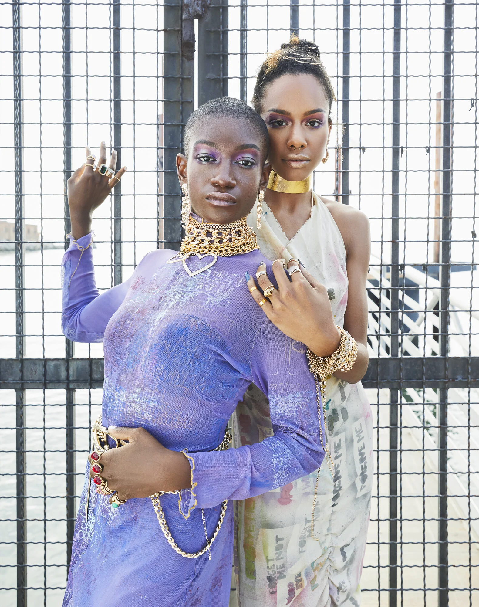 Two women with bold jewelry and colorful makeup posing against a metal fence with water and buildings in the background.