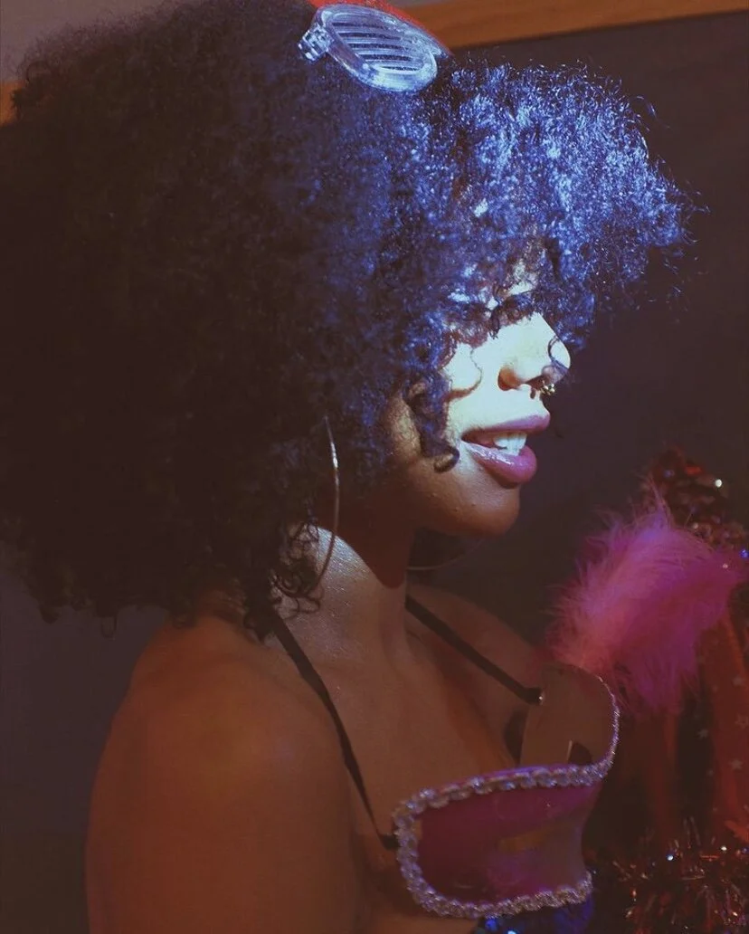 A woman with curly hair wearing glasses, earrings, and a pink top with embellishments, looking to the side in a dimly lit environment.