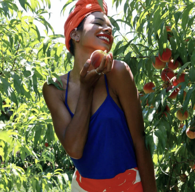 A woman with a red headscarf and red lipstick holding a peach, standing among peach trees with ripe peaches.