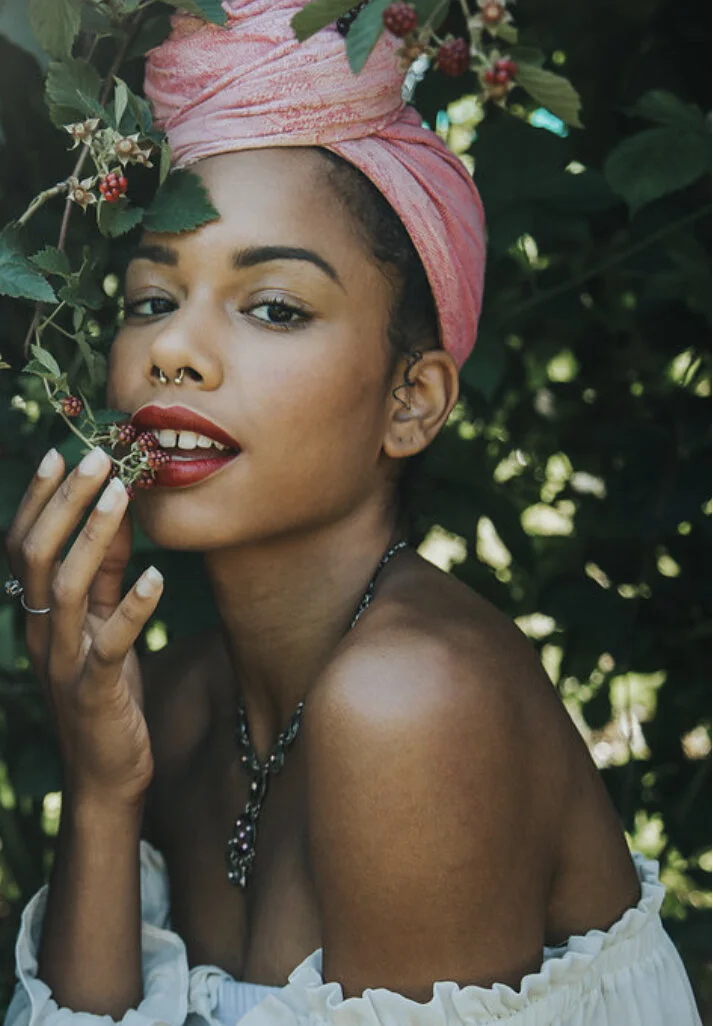 A woman with dark curly hair, wearing a pink headwrap, red lipstick, and a nose ring, is surrounded by green foliage and small red berries. She poses with a playful expression, touching berries near her lips, and wears a white off-shoulder top and a 