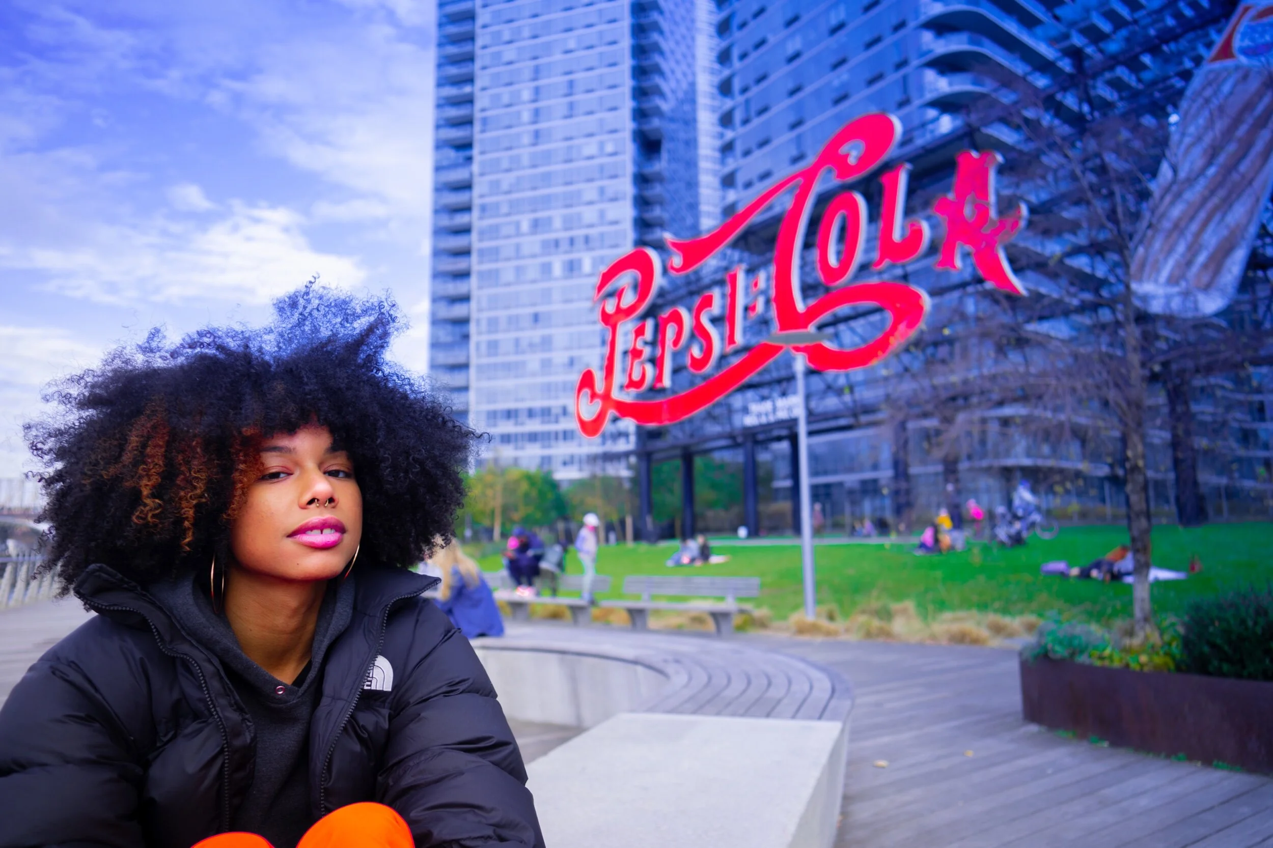 Young woman with curly hair and pink lipstick sitting outside in a park with high-rise buildings and a neon 'LOST' sign in the background.