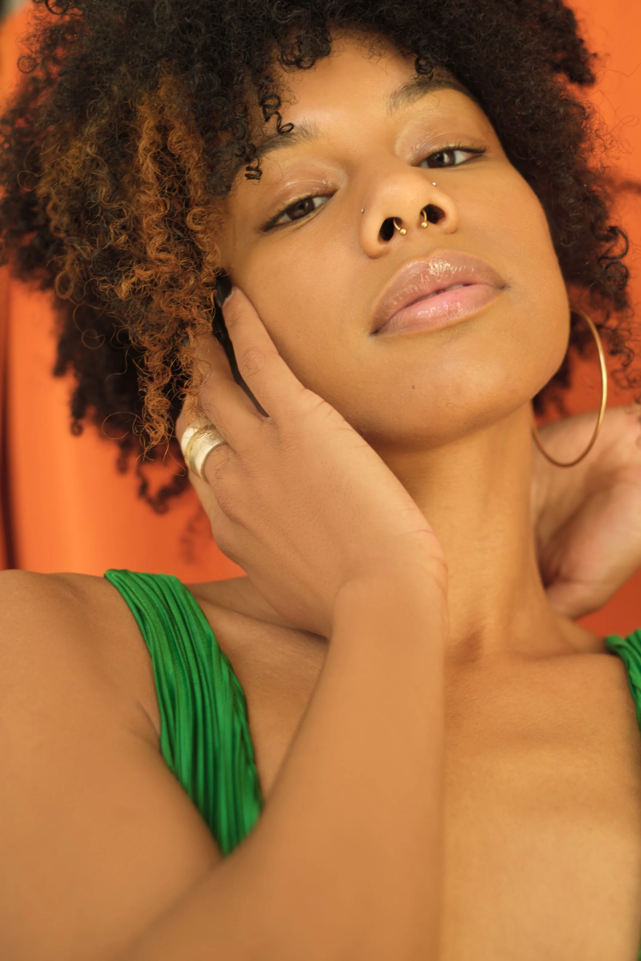 Close-up portrait of a woman with curly hair, wearing a green dress, gold hoop earrings, and a septum nose ring, touching her face, with an orange background.