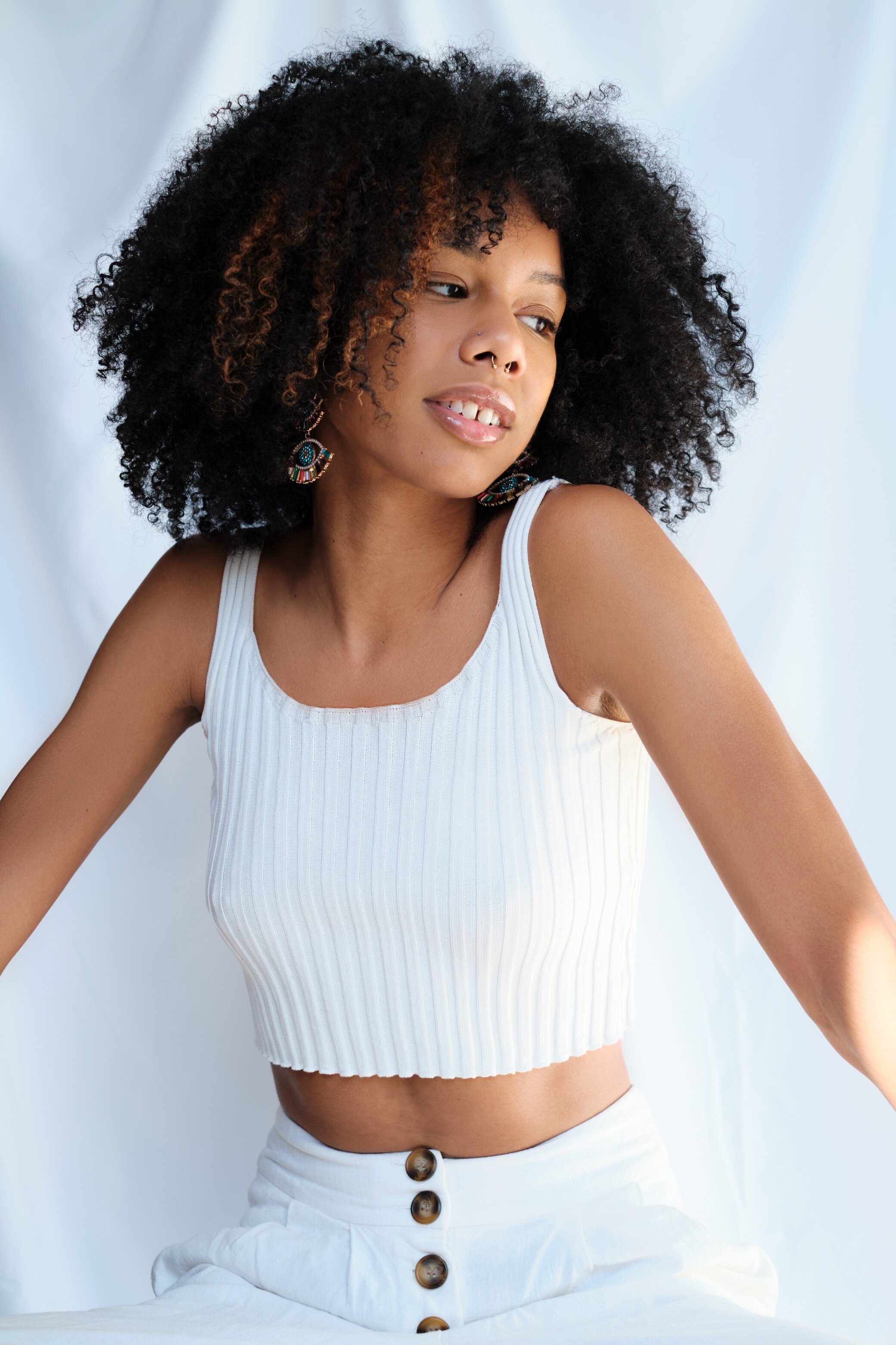 A young woman with curly black hair and wearing earrings looks off to the side, wearing a white sleeveless top and high-waisted white pants with brown buttons, against a white background.
