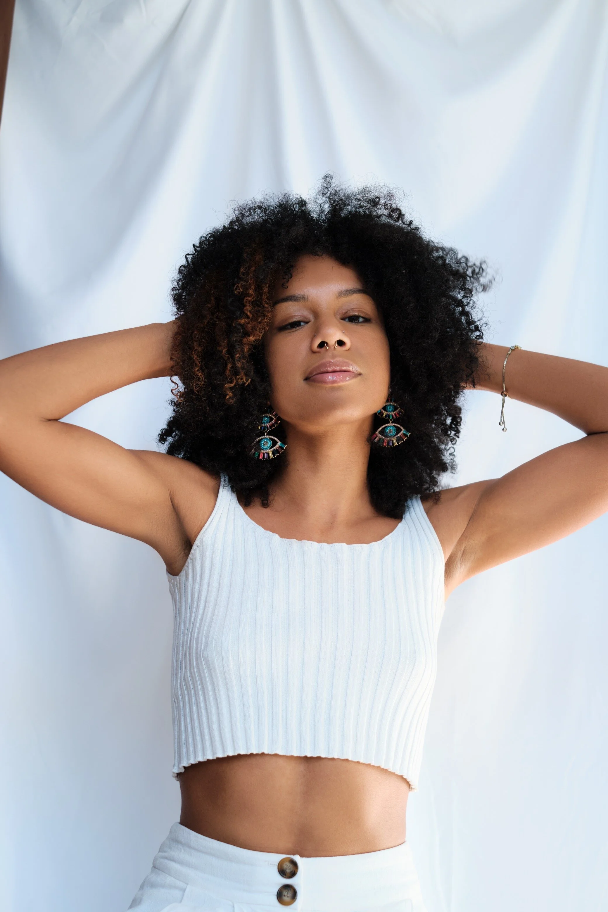 A young woman with curly hair, wearing a white ribbed crop top and high-waisted white pants, standing against a white backdrop, with her hands behind her head, looking directly at the camera with a confident expression.