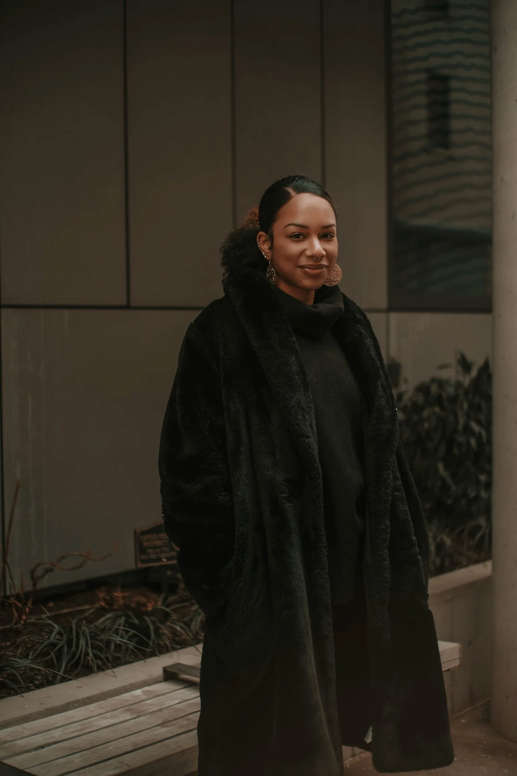 A woman with dark hair pulled back, wearing a black fur coat and large earrings, standing outside near a modern building with gray panels and some plants.