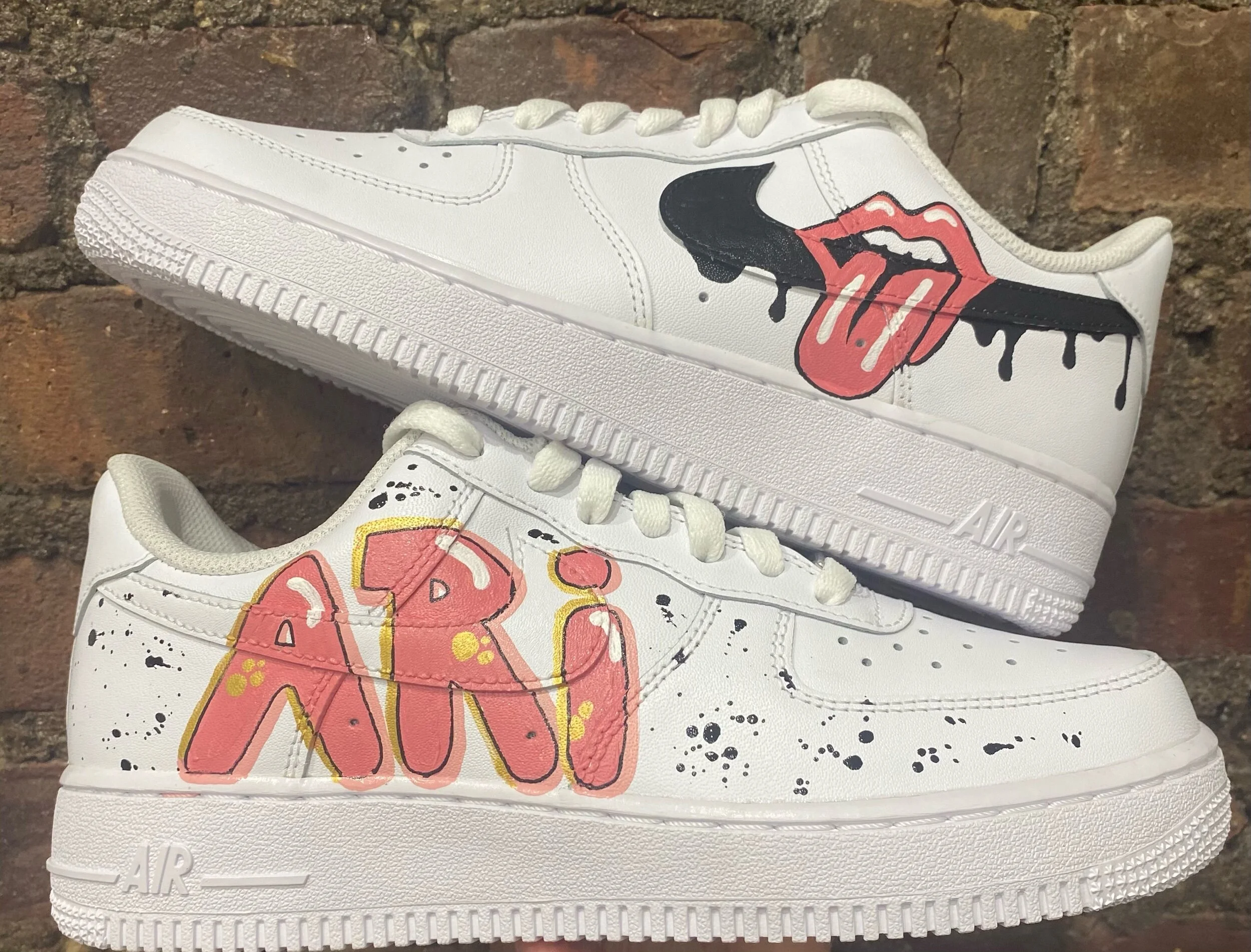 Close-up of a white sneaker with graffiti-style artwork, including a red tongue, black dripping paint, and abstract yellow and black designs, against a brick wall background.