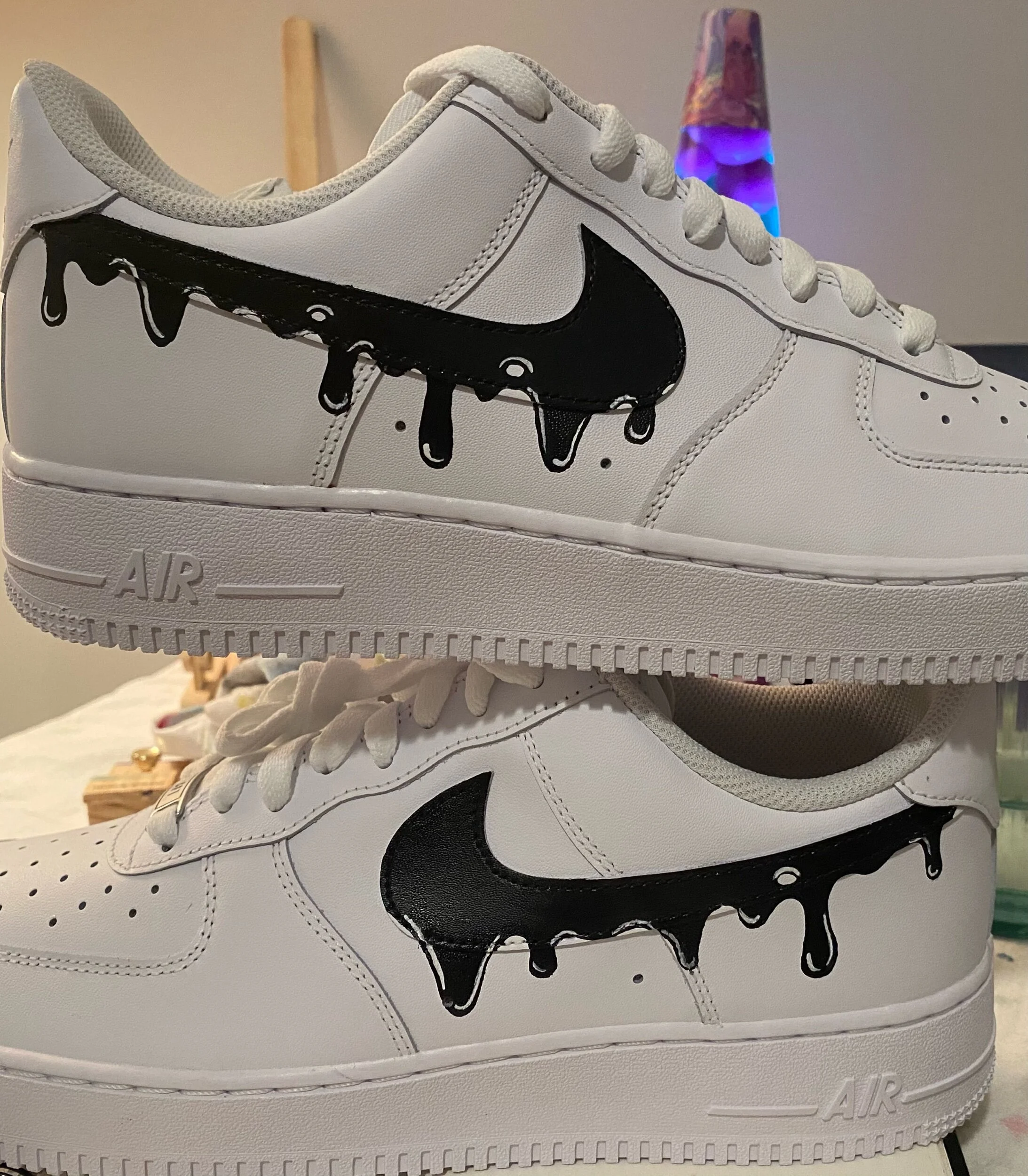 White sneakers with black dripping Nike swoosh logo and black paint drips on the side, on a white surface with a lava lamp in the background.
