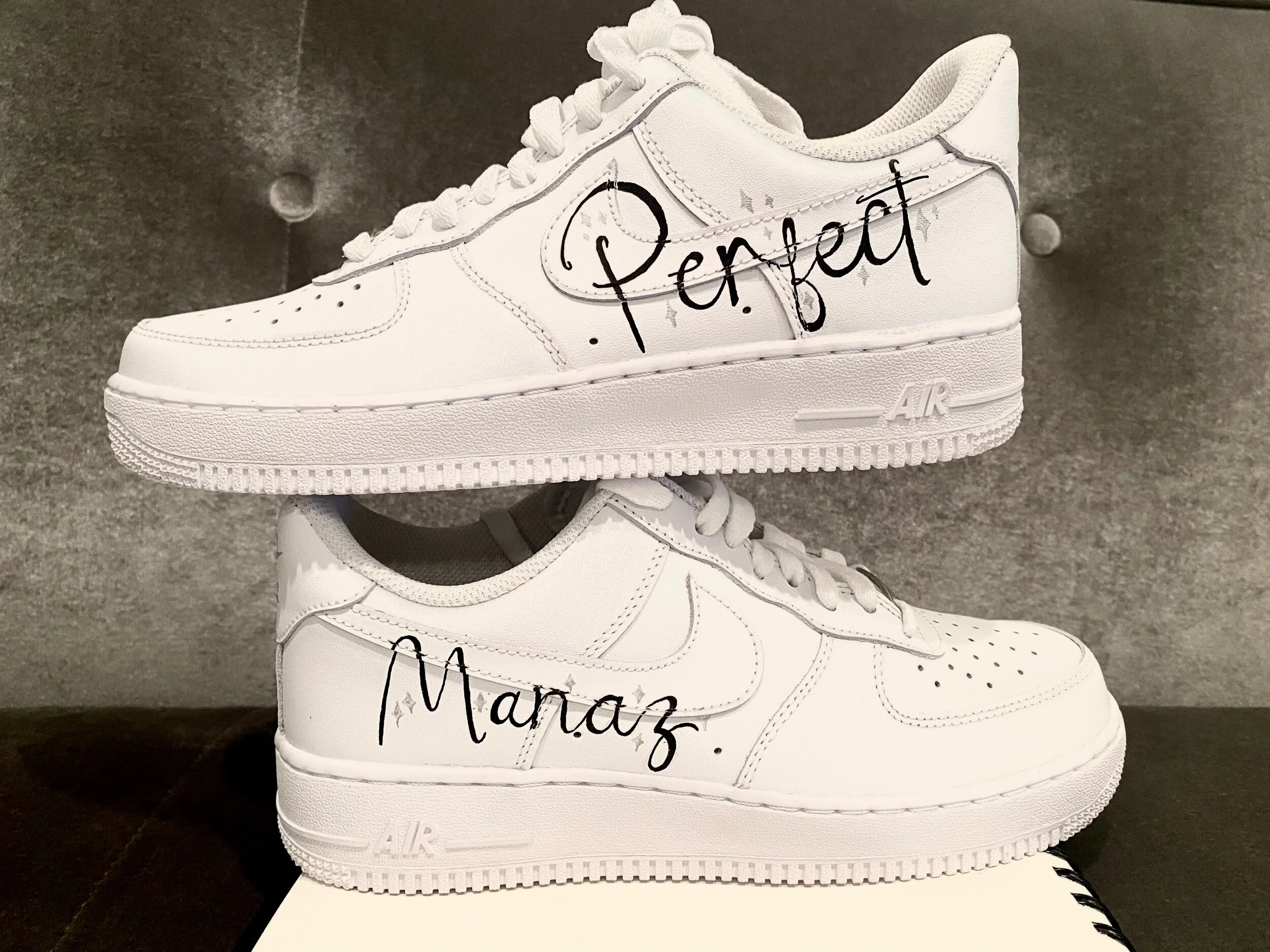 A white sneaker with black handwritten text and decorative stars on the side, placed on top of another similar sneaker, against a dark textured background.