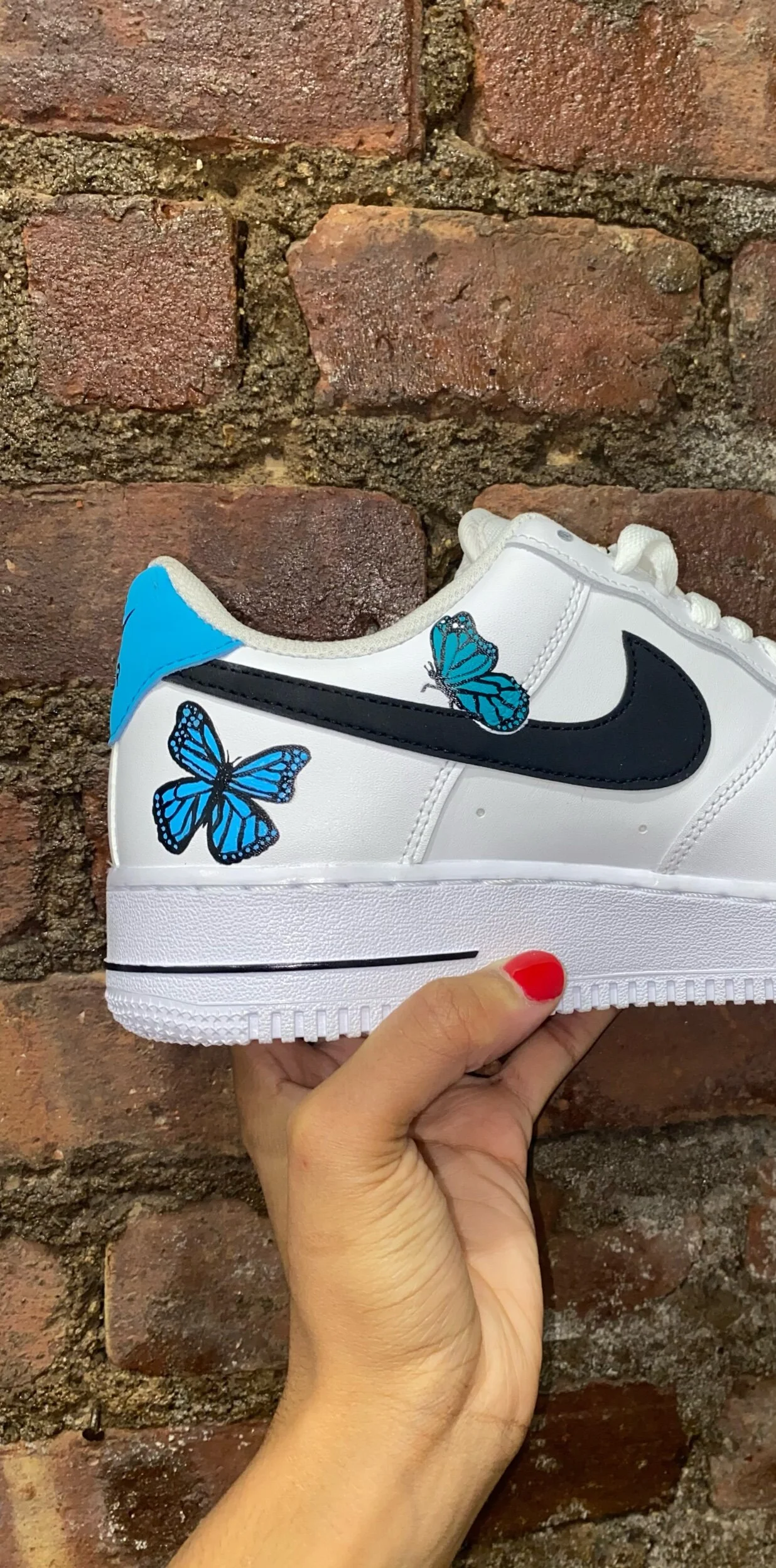 A white sneaker with black Nike swoosh logo, decorated with blue butterfly illustrations, held against a brick wall background.