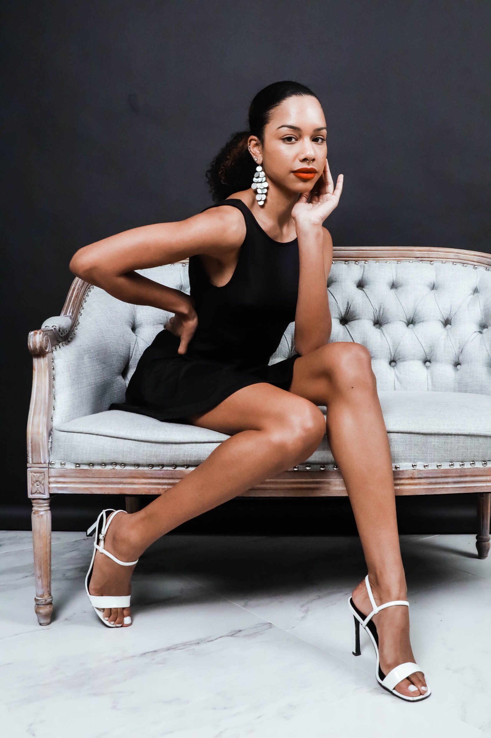 A woman with dark hair in a ponytail, wearing bold red lipstick, white strappy high heels, and statement earrings, sitting on an ornate beige and white vintage sofa against a dark wall, striking a pose with her right hand cupping her face.