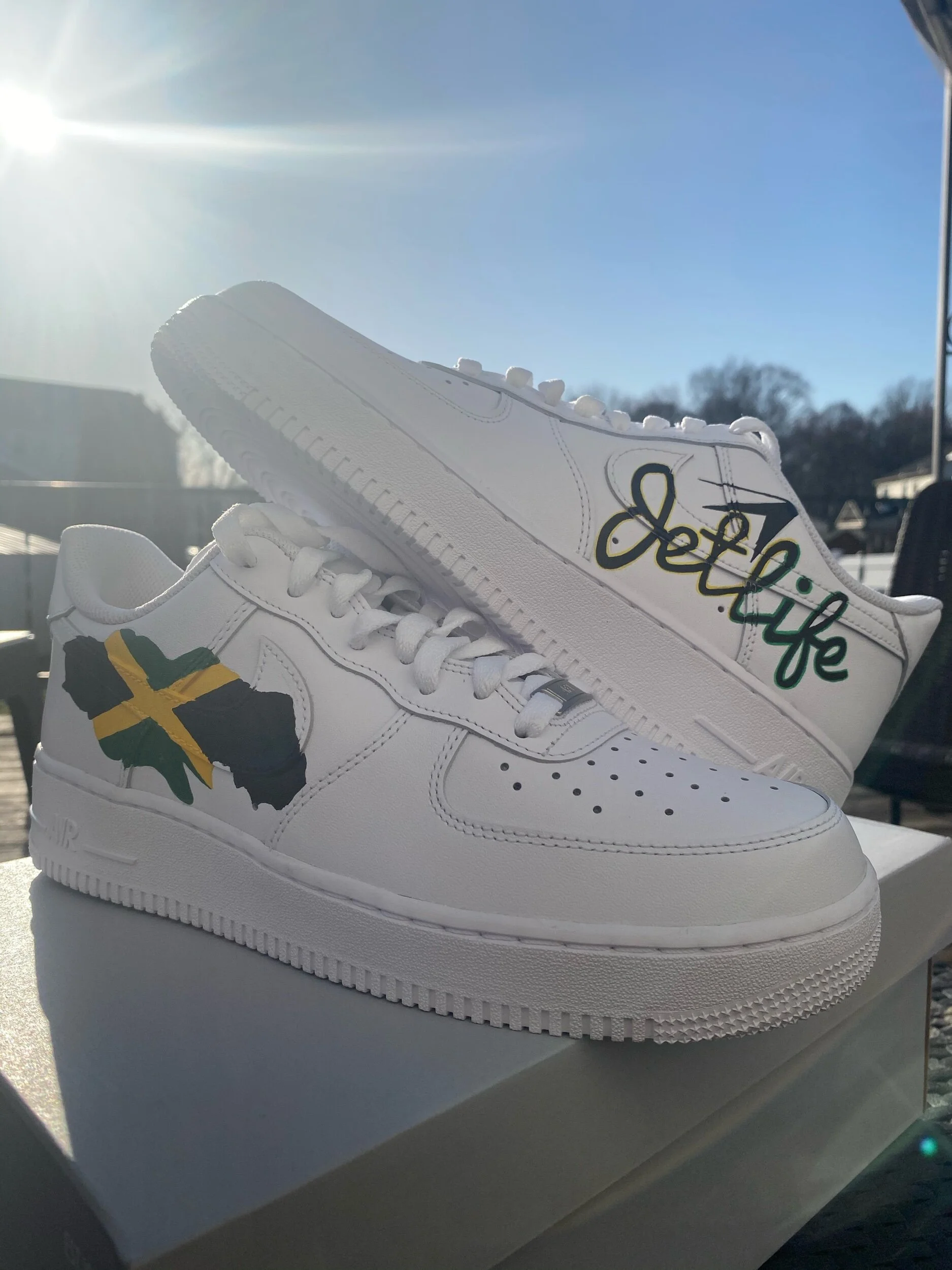 A pair of white Nike Air Force 1 sneakers with artistic signatures and designs on the side, placed on a white shoebox outside with a sunny sky in the background.