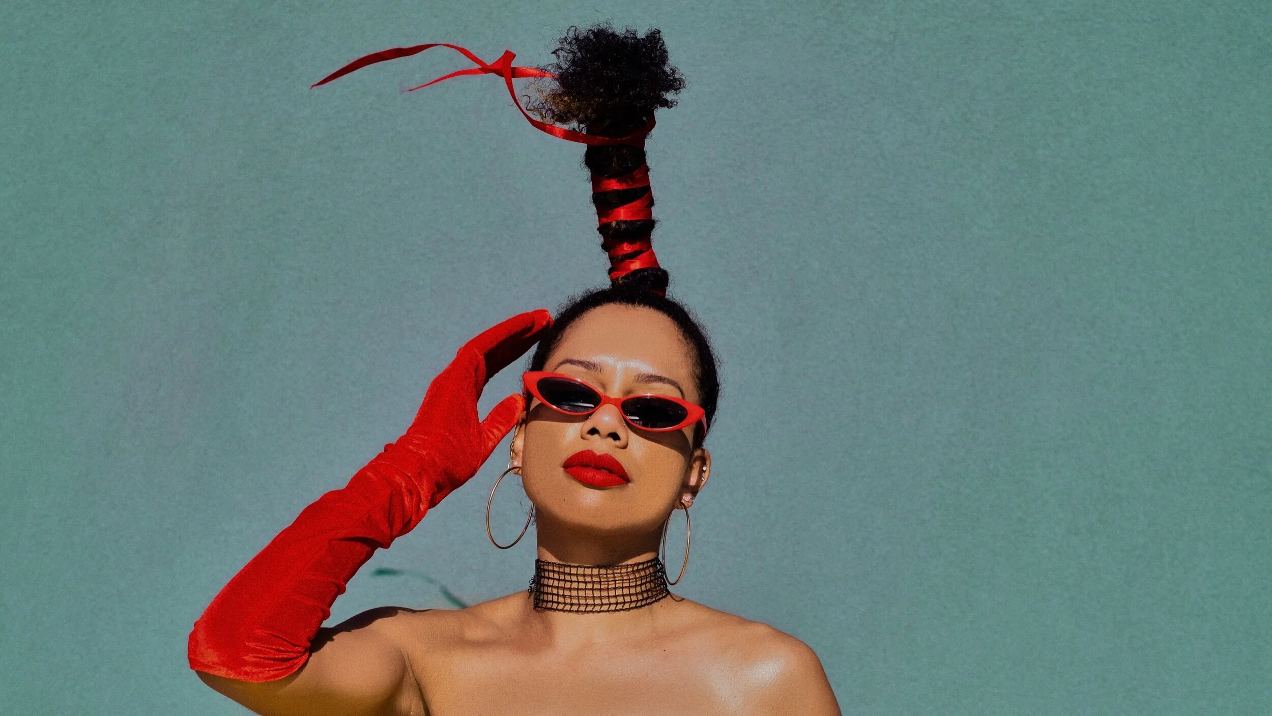 A woman with curly hair styled into a high ponytail wrapped with red ribbon, wearing red sunglasses, red gloves, hoop earrings, a black choker, and bold red lipstick, posing against a plain background.