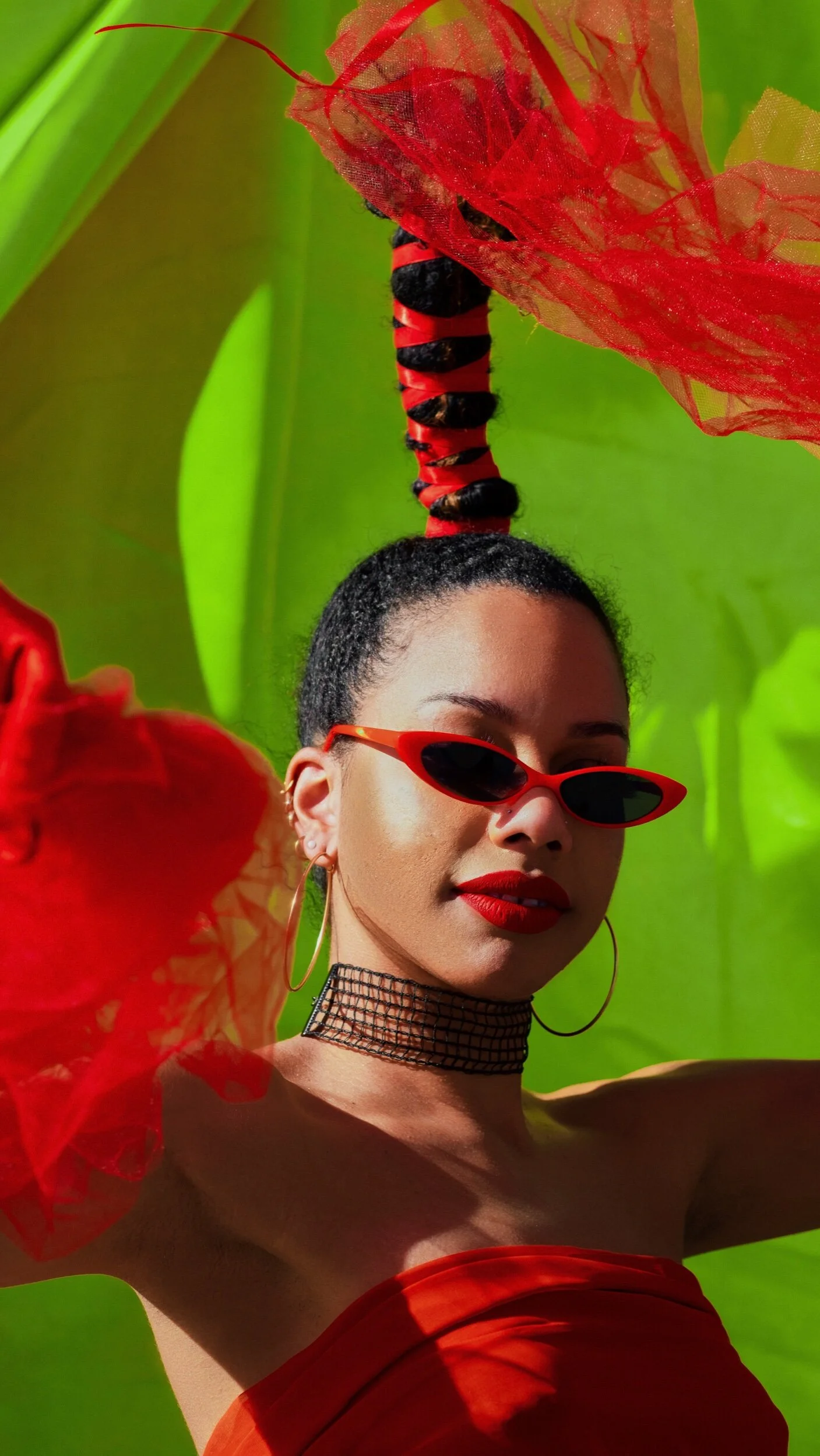 A woman with short curly dark hair, wearing red sunglasses, red lipstick, hoop earrings, and a black mesh choker, posing outdoors among large red flowers with a green background.