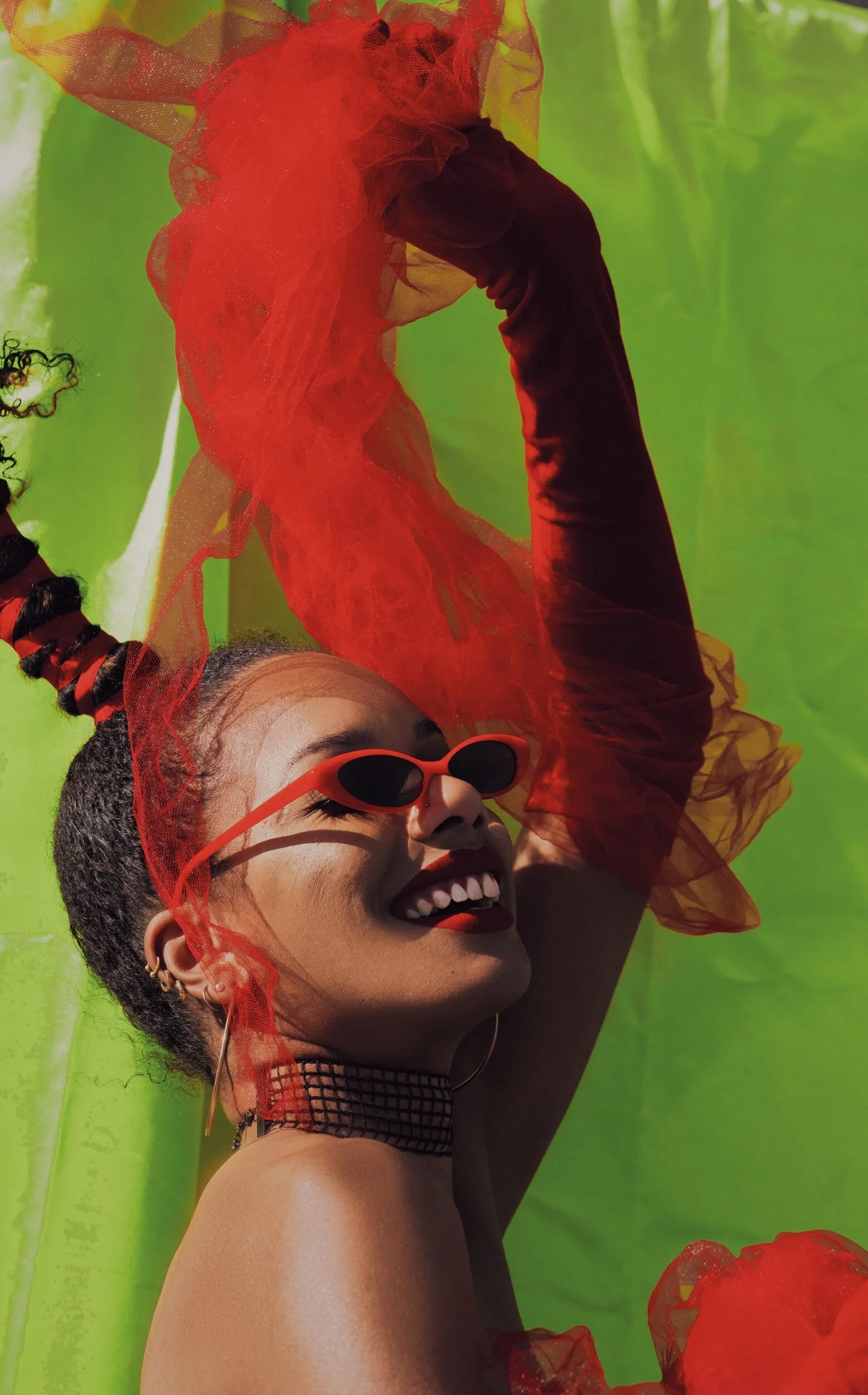 A smiling woman with short dark hair, wearing red sunglasses, a checkered choker, and earrings, surrounded by bright orange and red tulle fabric against a vibrant green background.