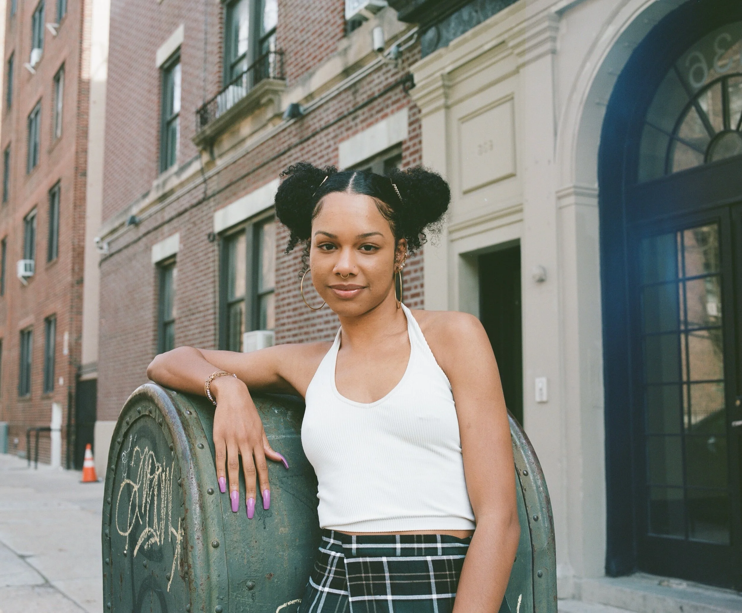 A young woman with curly hair styled in two buns is standing outdoors in front of a brick building with windows and a glass door. She is wearing a white tank top, plaid skirt, gold hoop earrings, and has purple fingernails. She is leaning on a green 