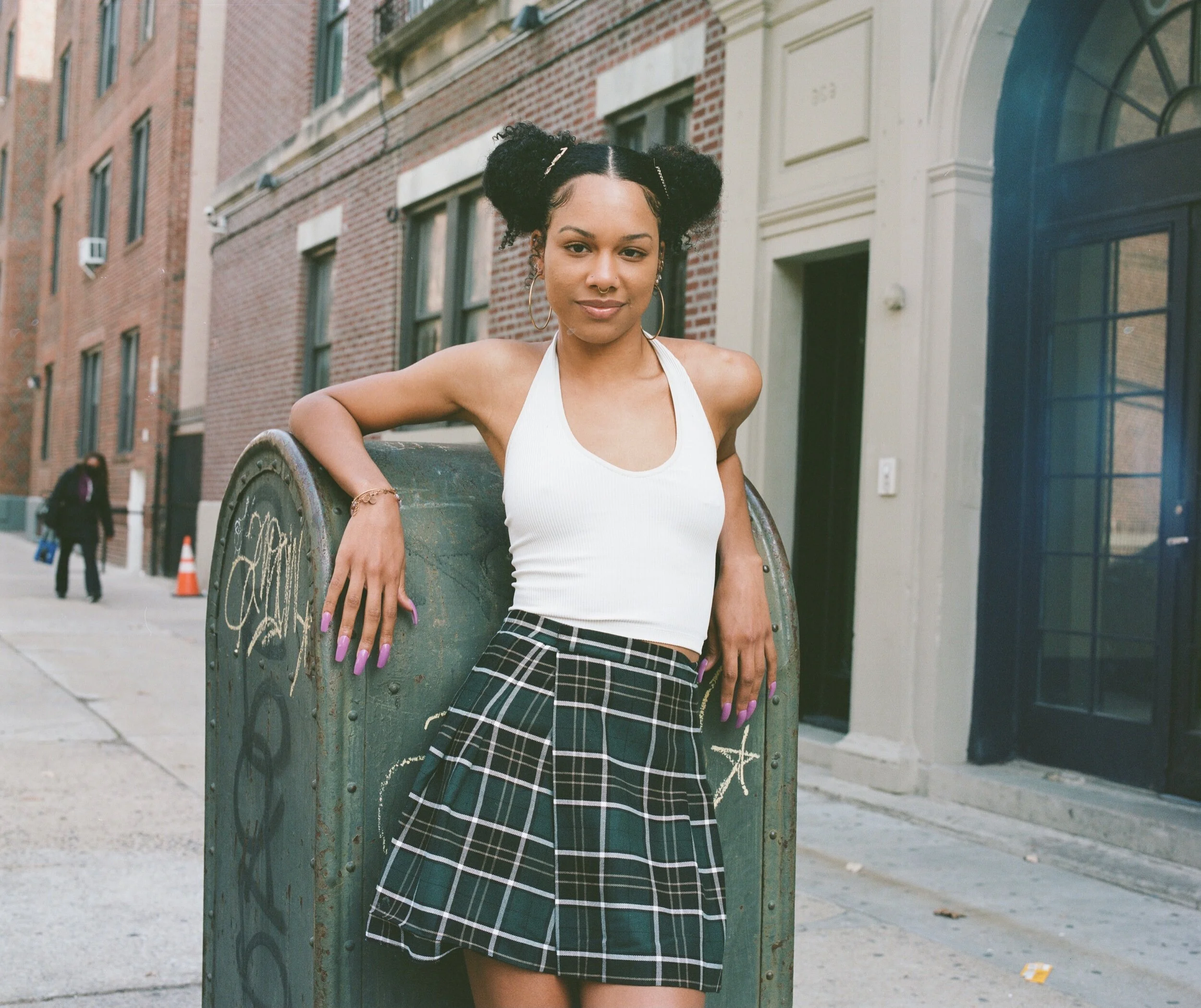 A young woman with dark curly hair styled in two buns, wearing a white tank top and a plaid skirt, standing outdoors in front of a brick building with windows. She leans on a decorated trash can, smiling at the camera.