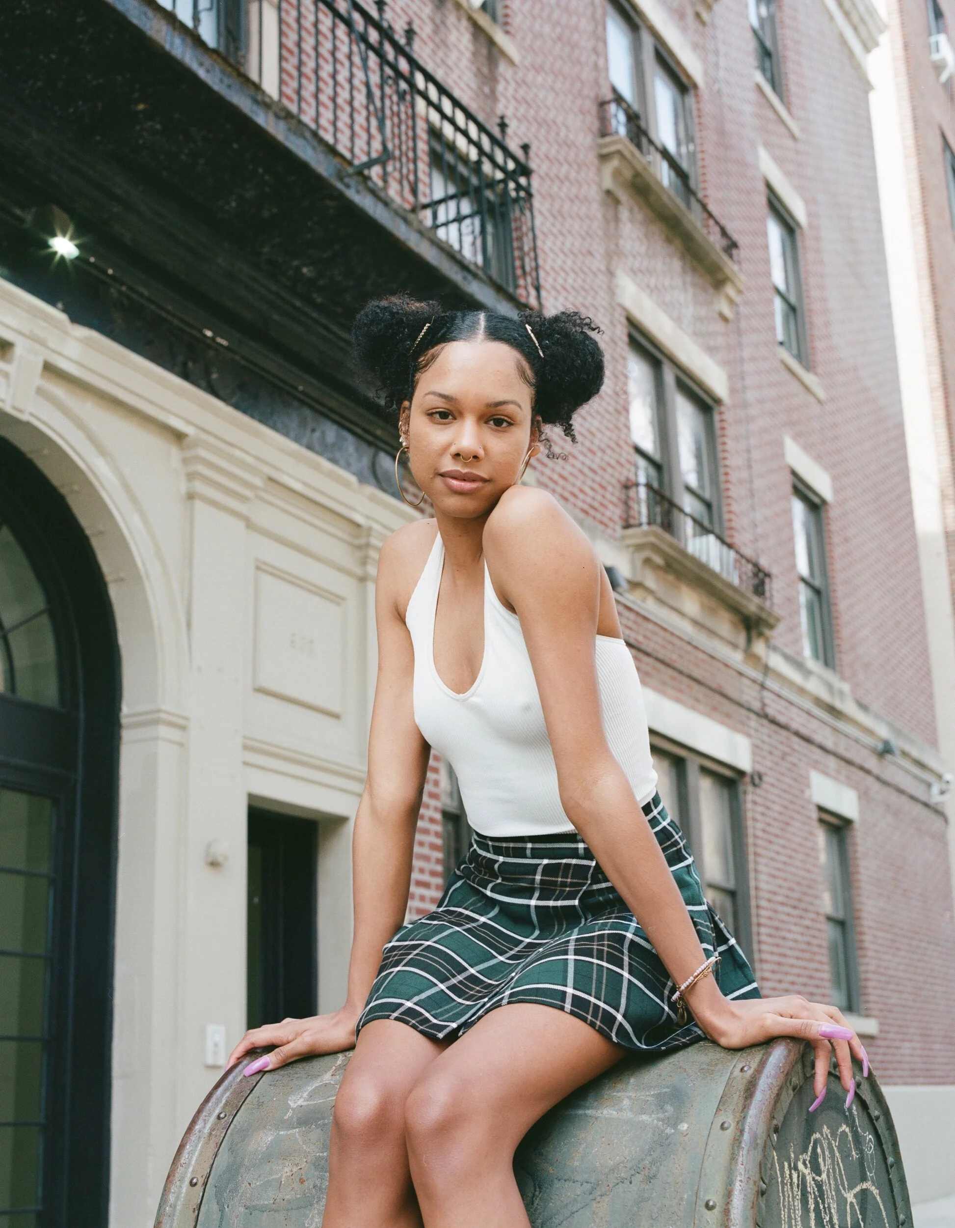 A young woman with curly hair styled in two buns, wearing a white tank top and a plaid skirt, sits on a metal barrel outside in an urban environment with red brick buildings and black balconies in the background.
